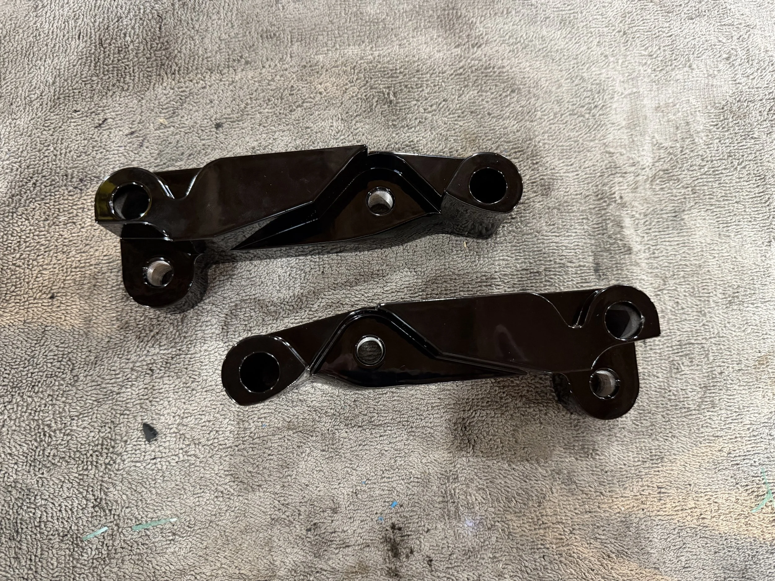 Two black metal motorcycle parts with mounting holes placed on a textured beige cloth.