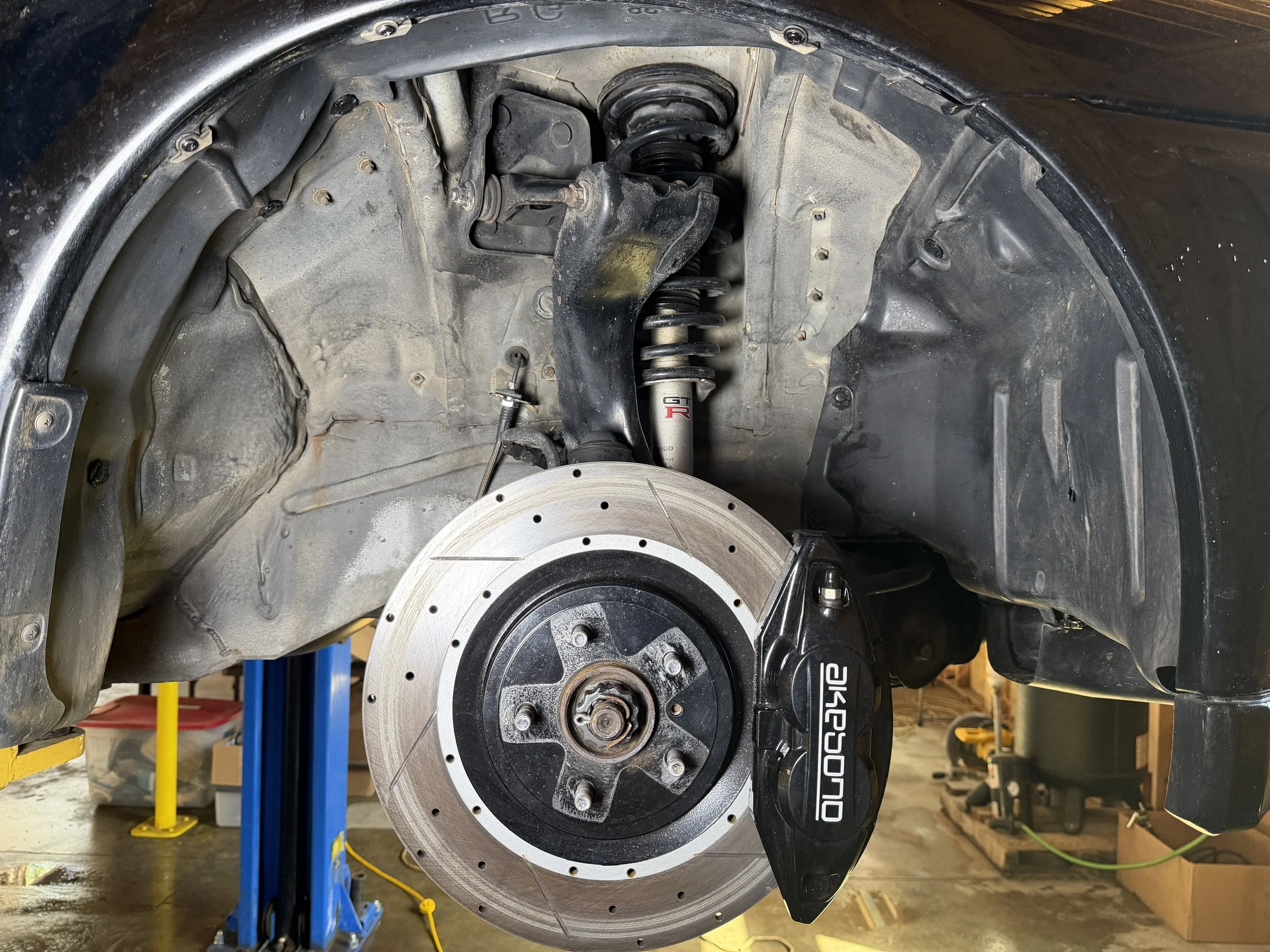 Close-up of a car's front suspension and brake system showing coil spring, shock absorber, brake caliper labeled 'Okuyama', and drilled brake disc, with part of the wheel removed.