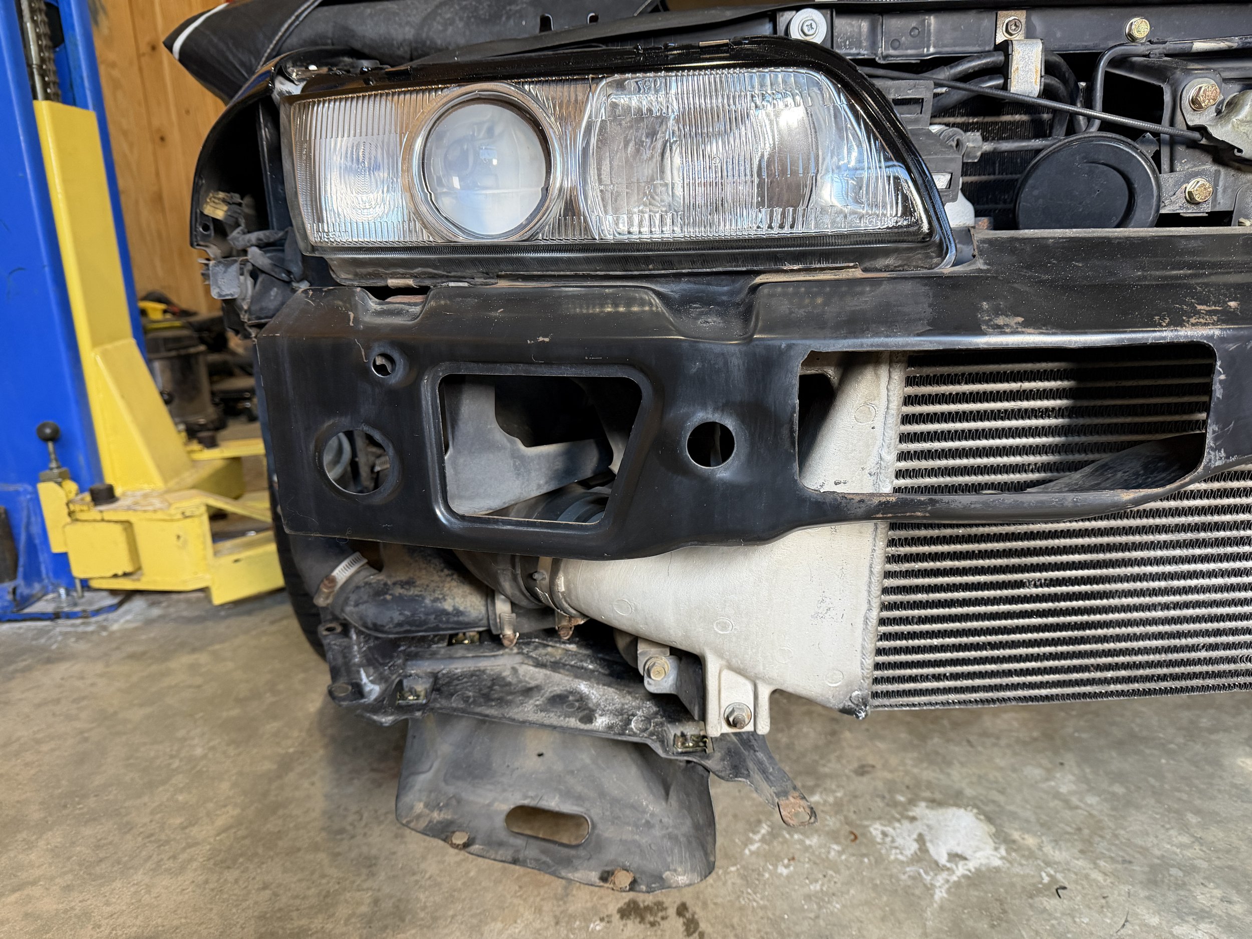 Close-up of a black car front end with missing bumper, showing headlight, radiator, and engine components, on a concrete floor in a garage.