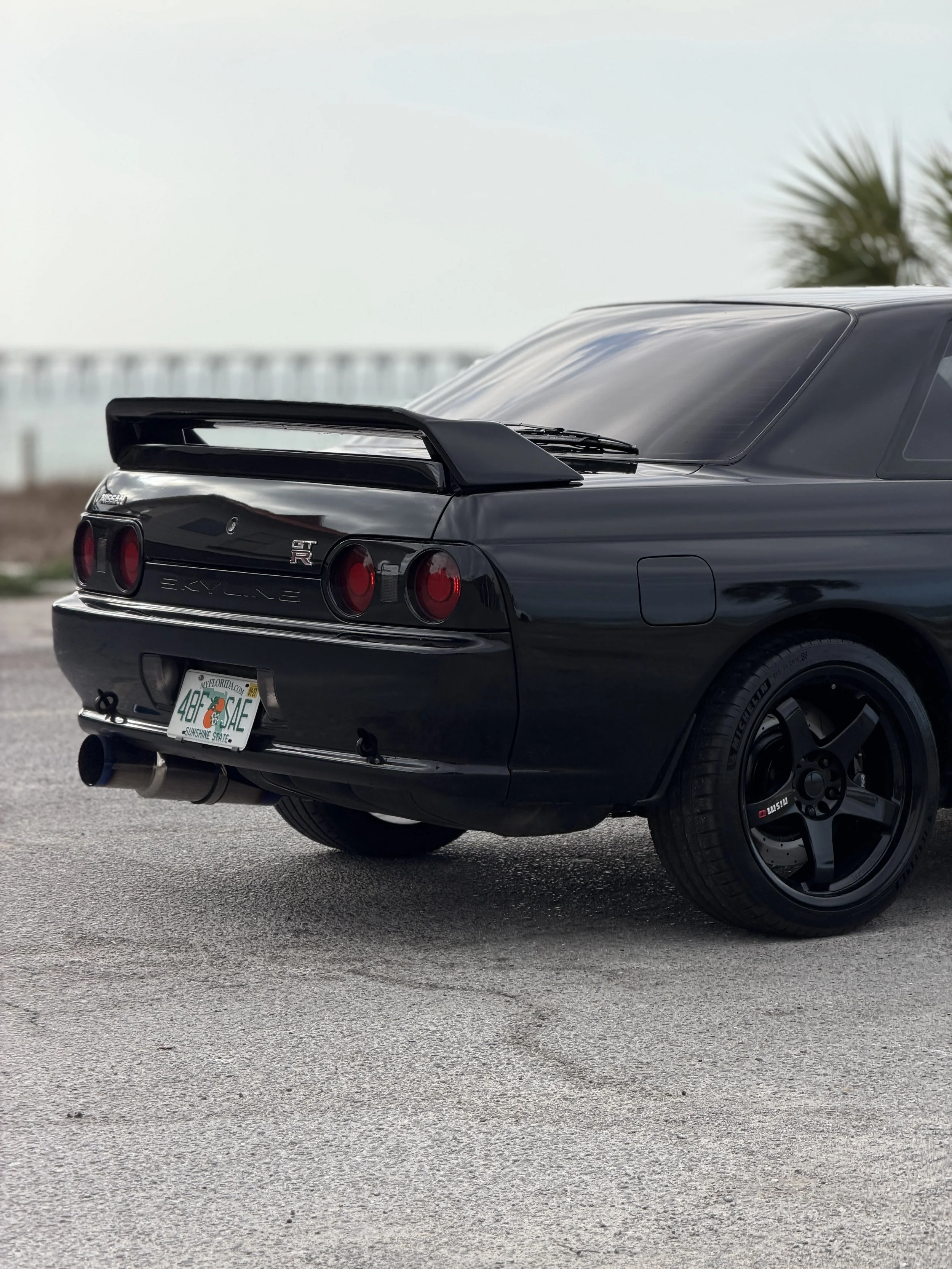 R32 NISSAN SKYLINE GTR FOR SALE
Black Nissan Skyline GT-R parked on a road with a blurred Ocean and palm tree in the background.