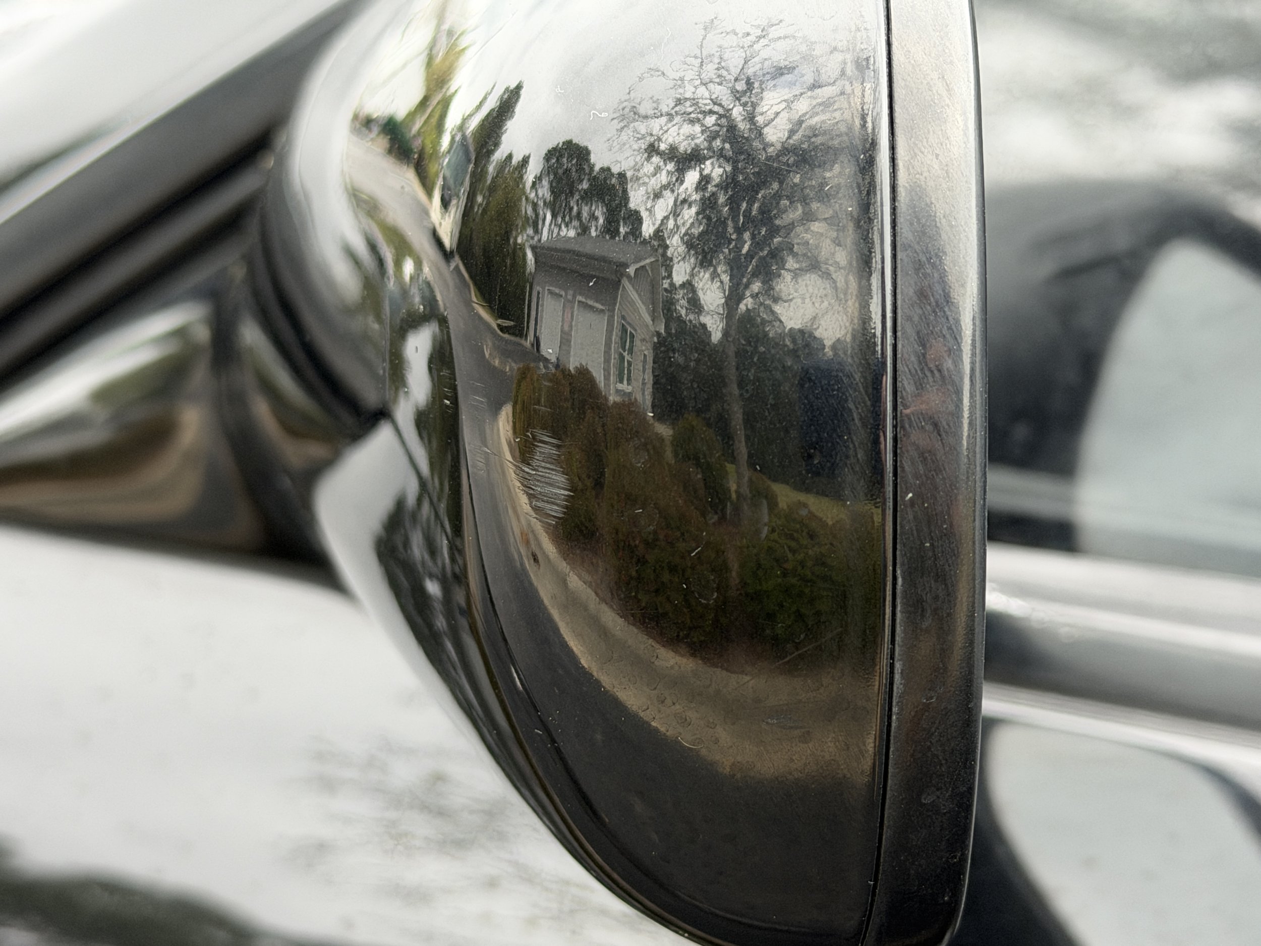 Close-up of a black car side mirror reflecting a house, trees, and an overcast sky.