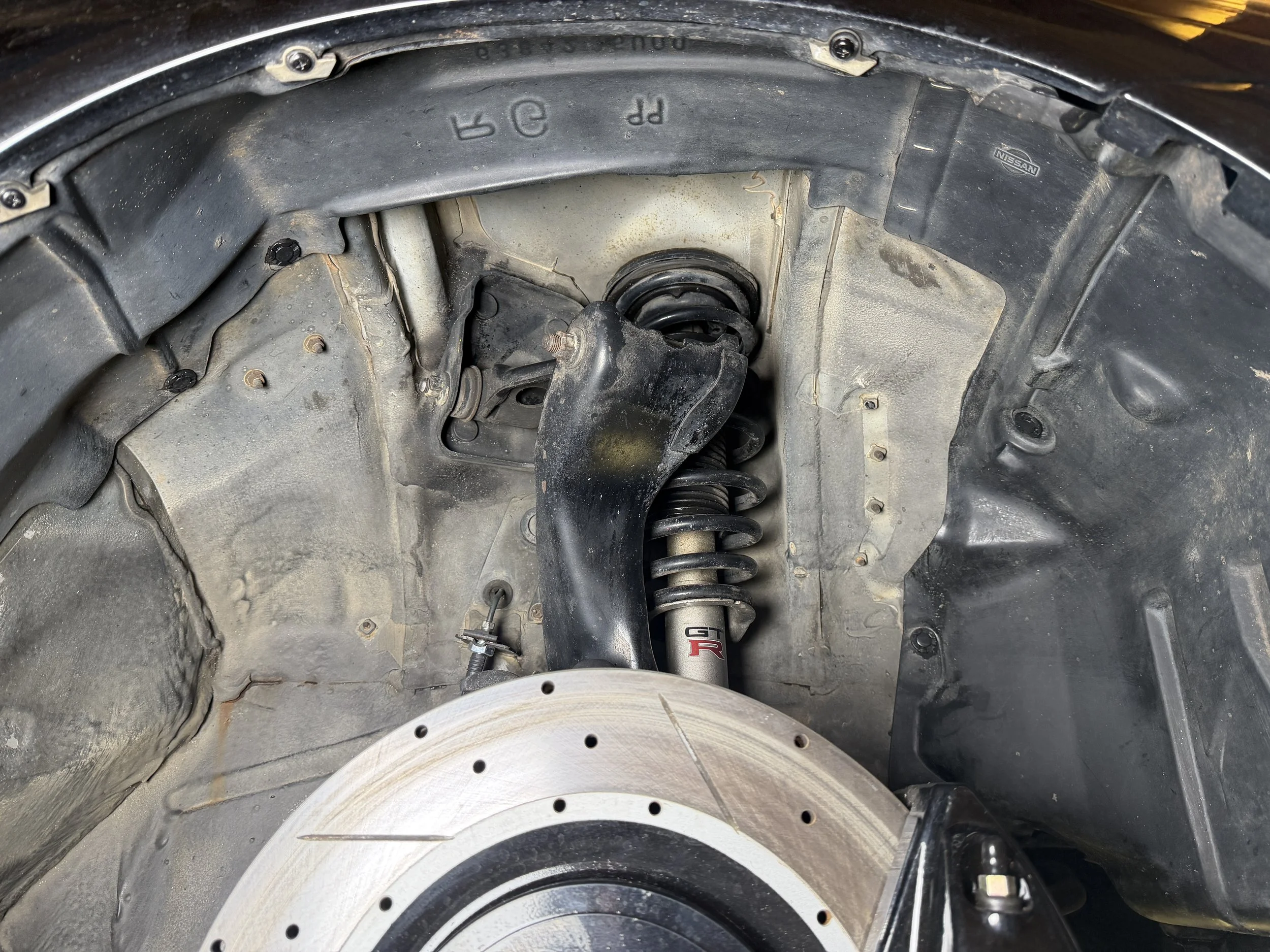 Close-up of a car's wheel well showing a suspension coil spring and shock absorber, with a brake rotor partially visible at the bottom.