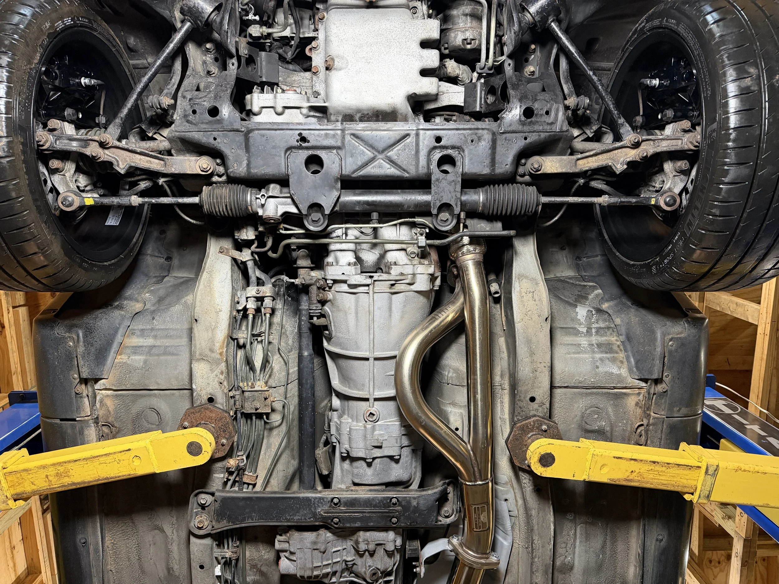 Underneath view of a car's engine, suspension, and exhaust system, supported by yellow lift arms in a garage.