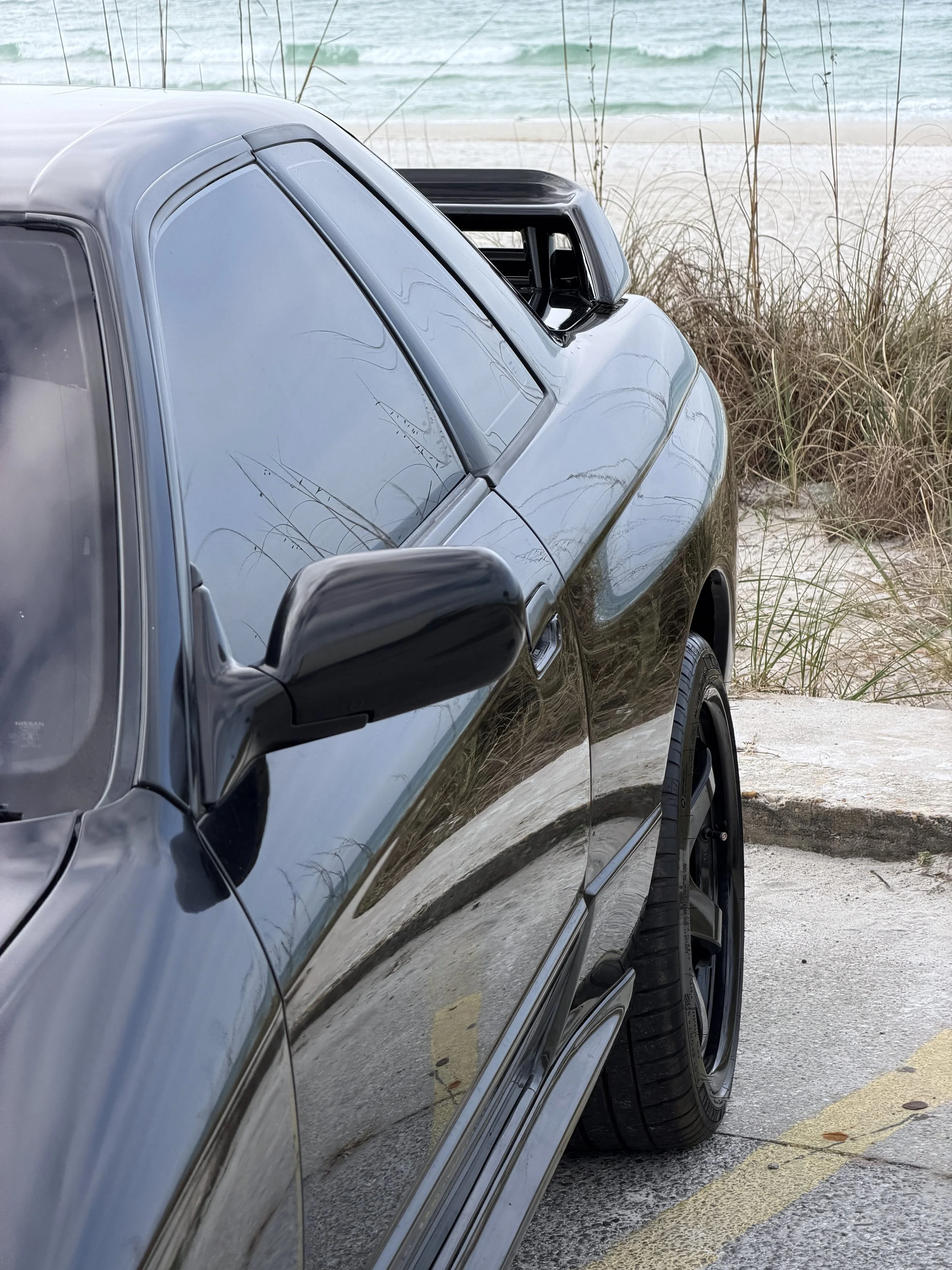 A black sports car parked on a sandy beach near the ocean with tall beach grass and gentle waves in the background.