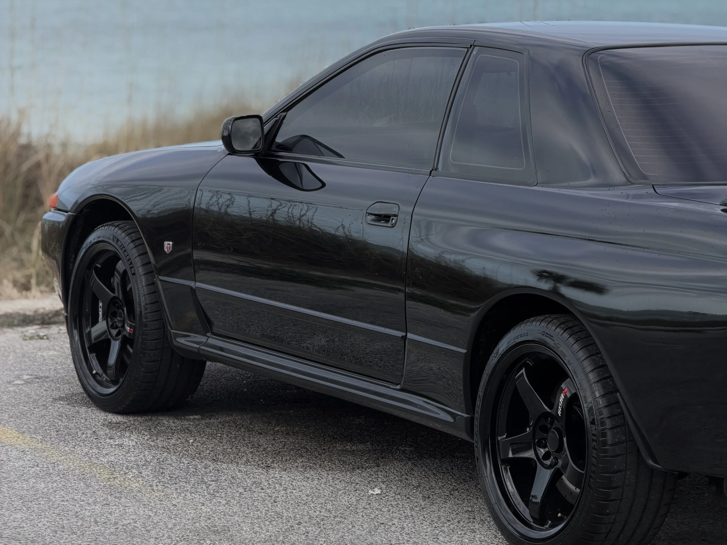 R32 NISSAN SKYLINE GTR FOR SALE
Black sports car parked on road with water and grass in background.