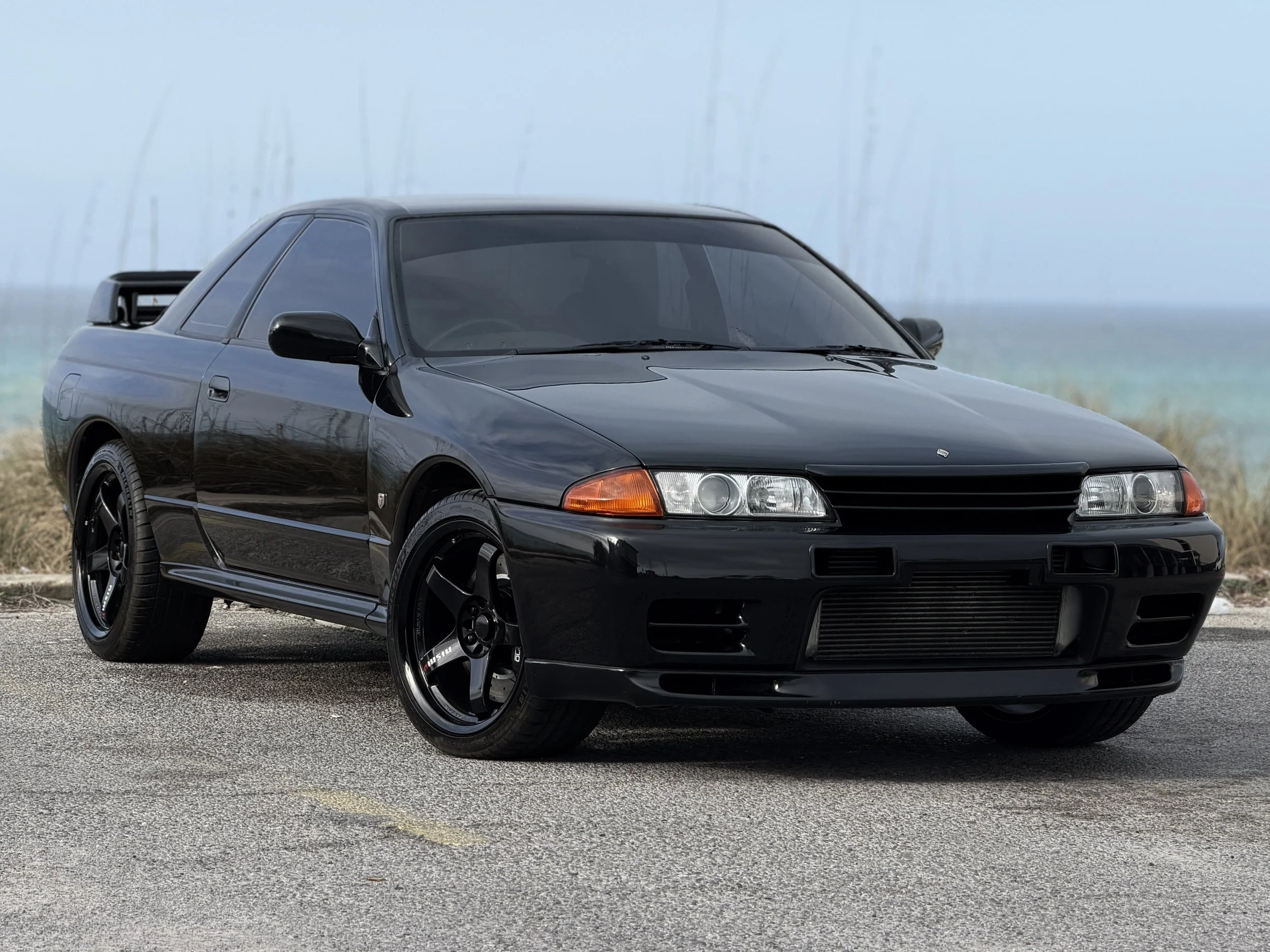R32 NISSAN SKYLINE GTR FOR SALE Black Nissan GTR sports car parked on a road near a beach with grassy dunes and water in the background.