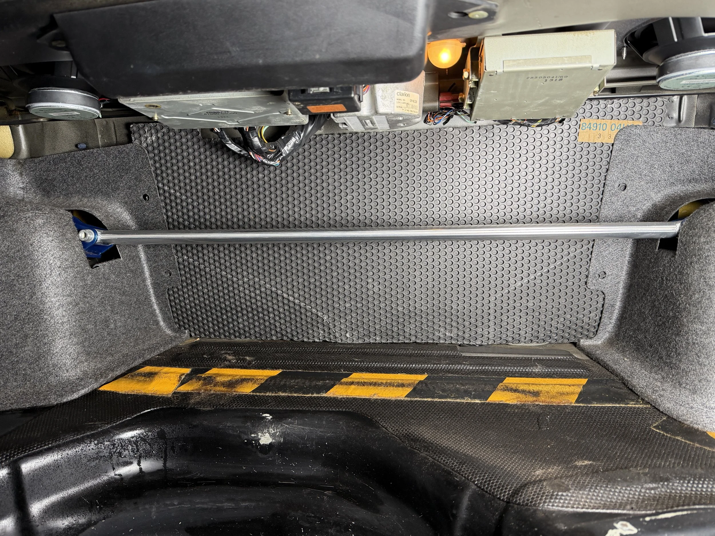 Interior of a vehicle with a grey floor mat, carpeted side panels, a metal support bar, and some electronic equipment underneath the dashboard.