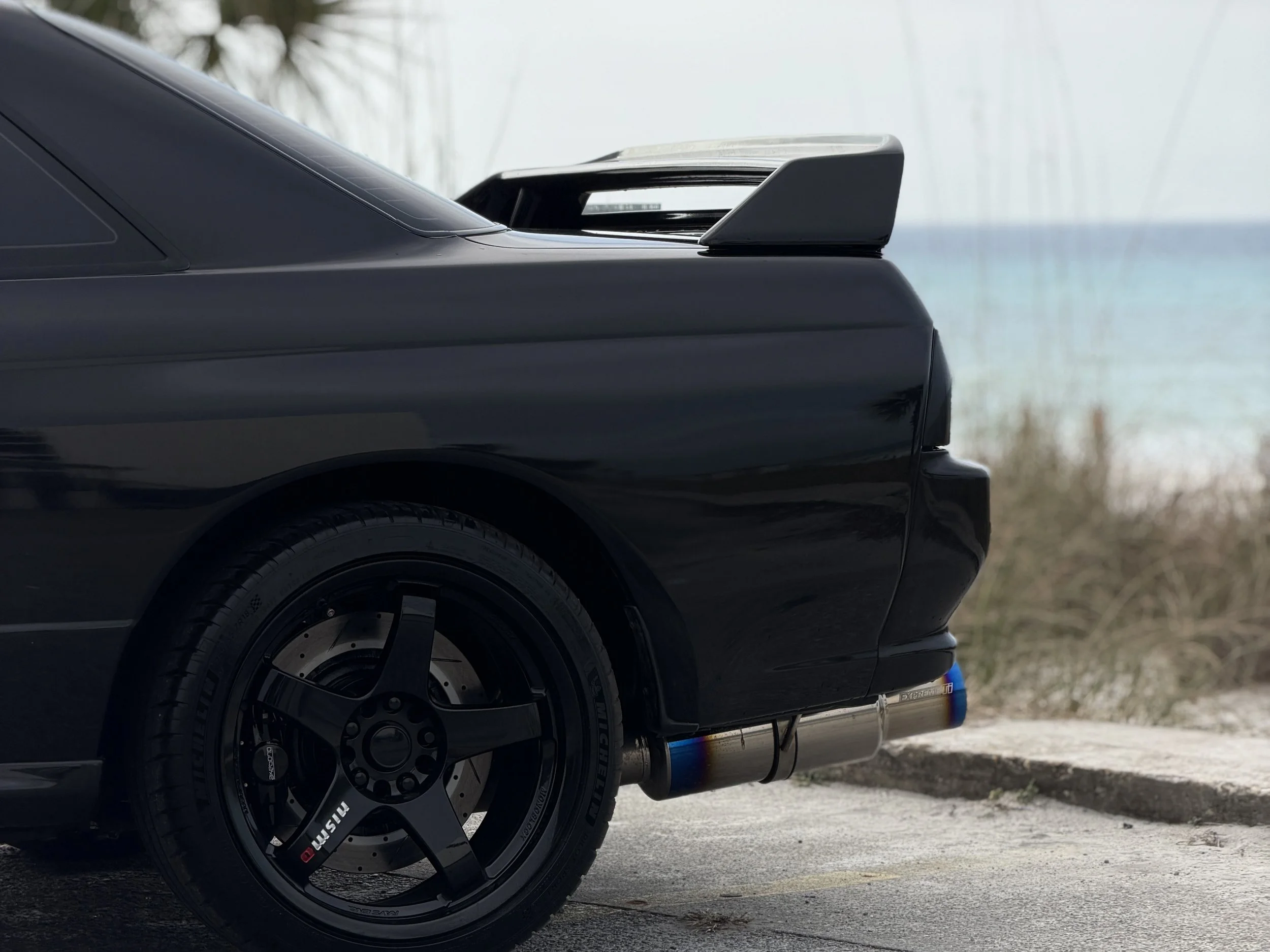 R32 NISSAN SKYLINE GTR FOR SALE
Close-up of the rear side of a black sports car with a spoiler, black wheel, and exhaust pipe, near a beach.