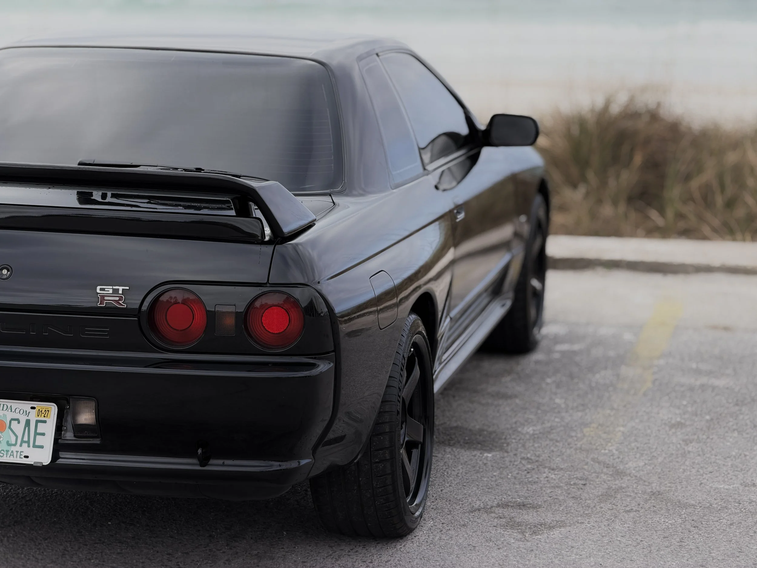 R32 NISSAN SKYLINE GTR FOR SALE
Rear view of a black Nissan Skyline GT-R sports car parked in an outdoor lot with grass and water in the background.
