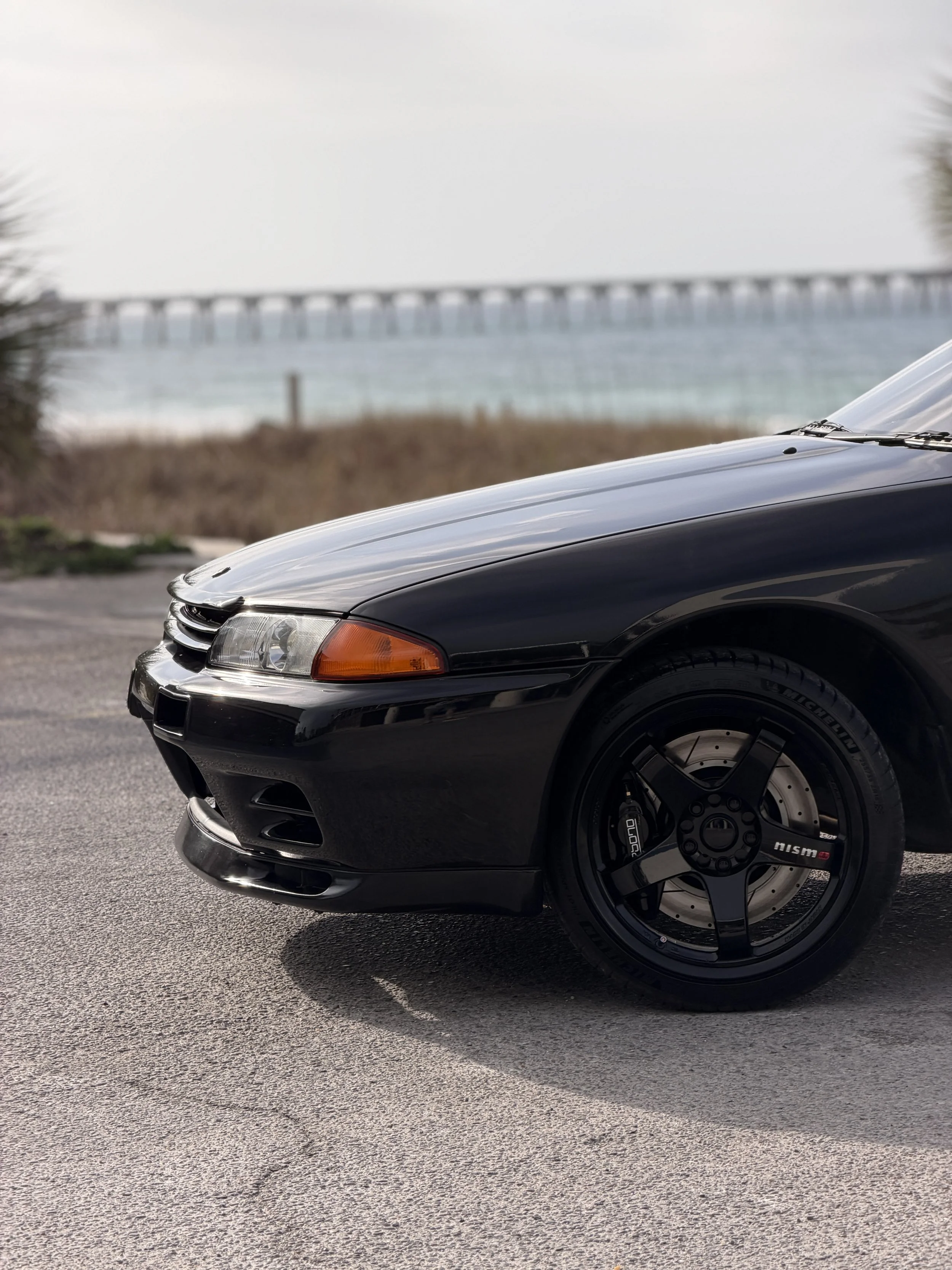 R32 NISSAN SKYLINE GTR FOR SALE
Close-up of the front side of a black sports car with black rims parked on a paved surface near a beach with a bridge in the background