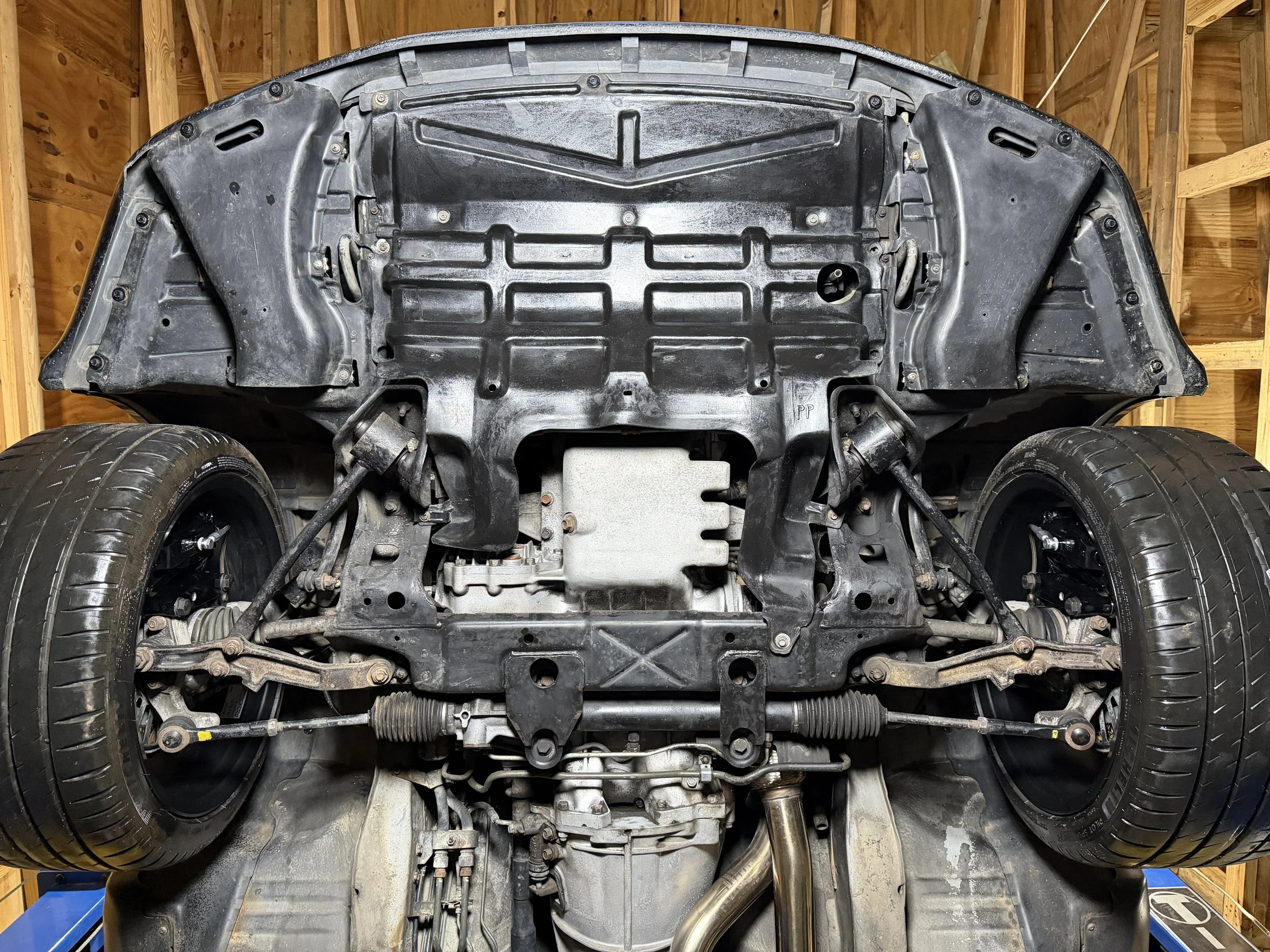 View of the underside of a car in a garage, showing the car's chassis, suspension components, wheels, and exhaust system.