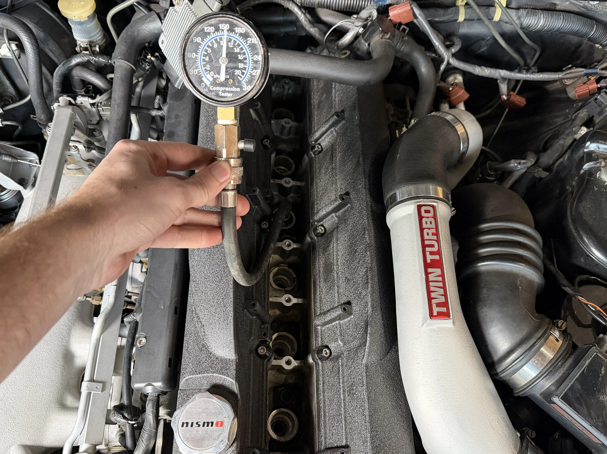 Person holding a pressure gauge to an engine, with a Twin Turbo intake pipe and various engine components visible.