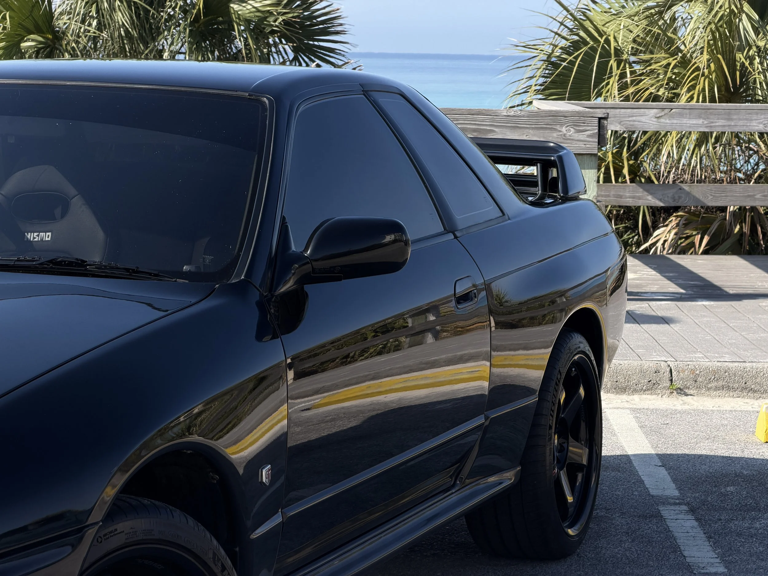 Close-up of a black car parked near the beach, with palm trees and ocean in the background.