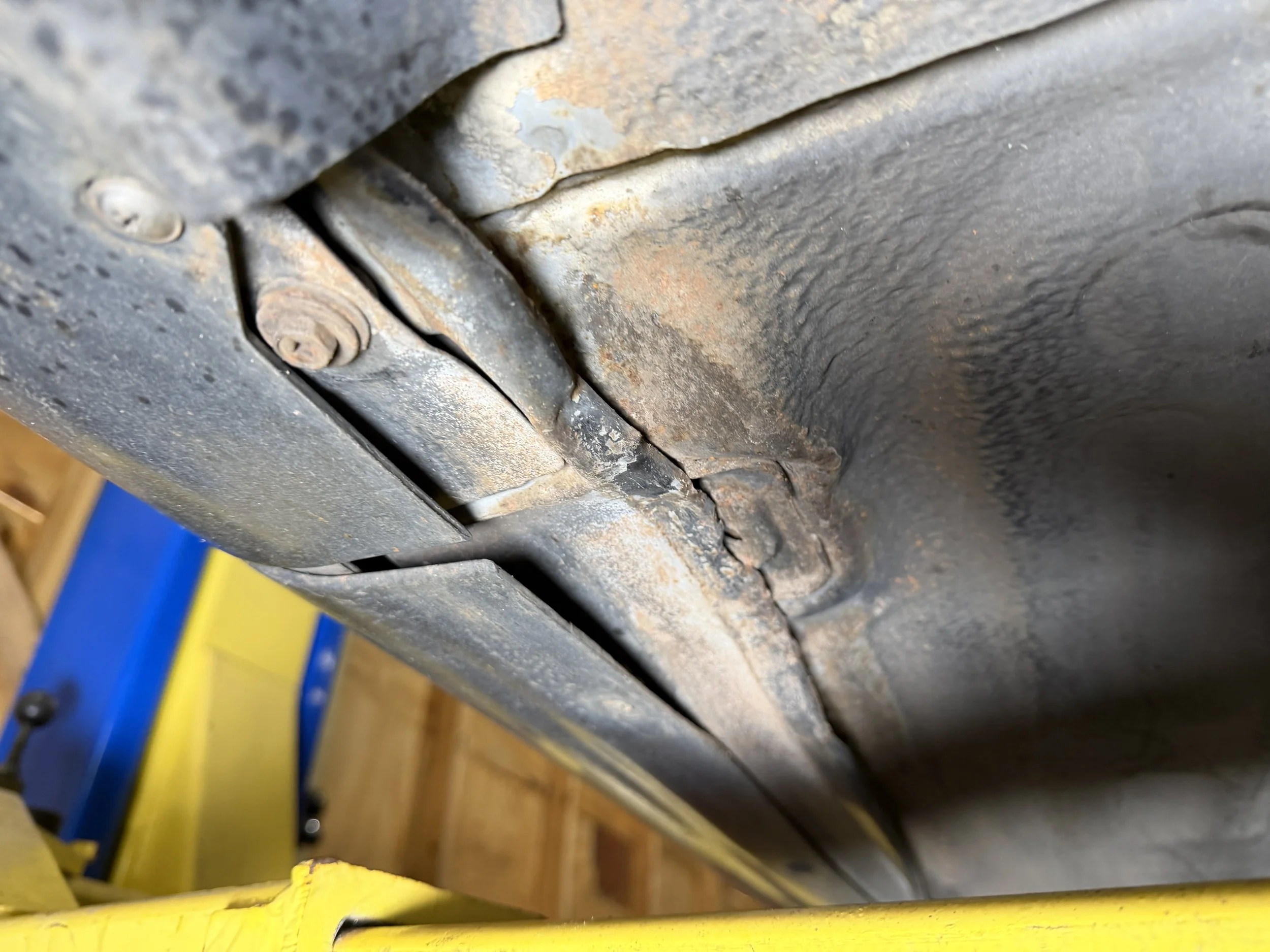 Underneath view of a rusty, old car frame with bolts and metal parts, supported on yellow and blue lift platform.