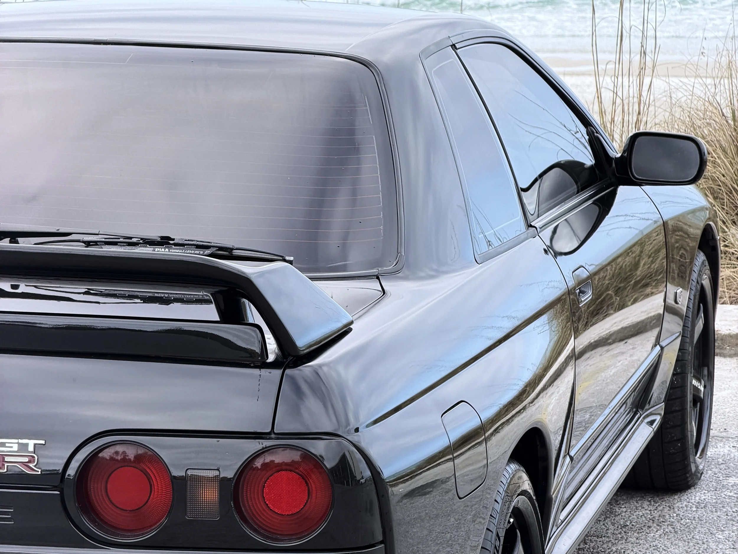 Close-up of a black Nissan GT-R sports car parked near a body of water with dry grass nearby, showing the rear and side of the vehicle.