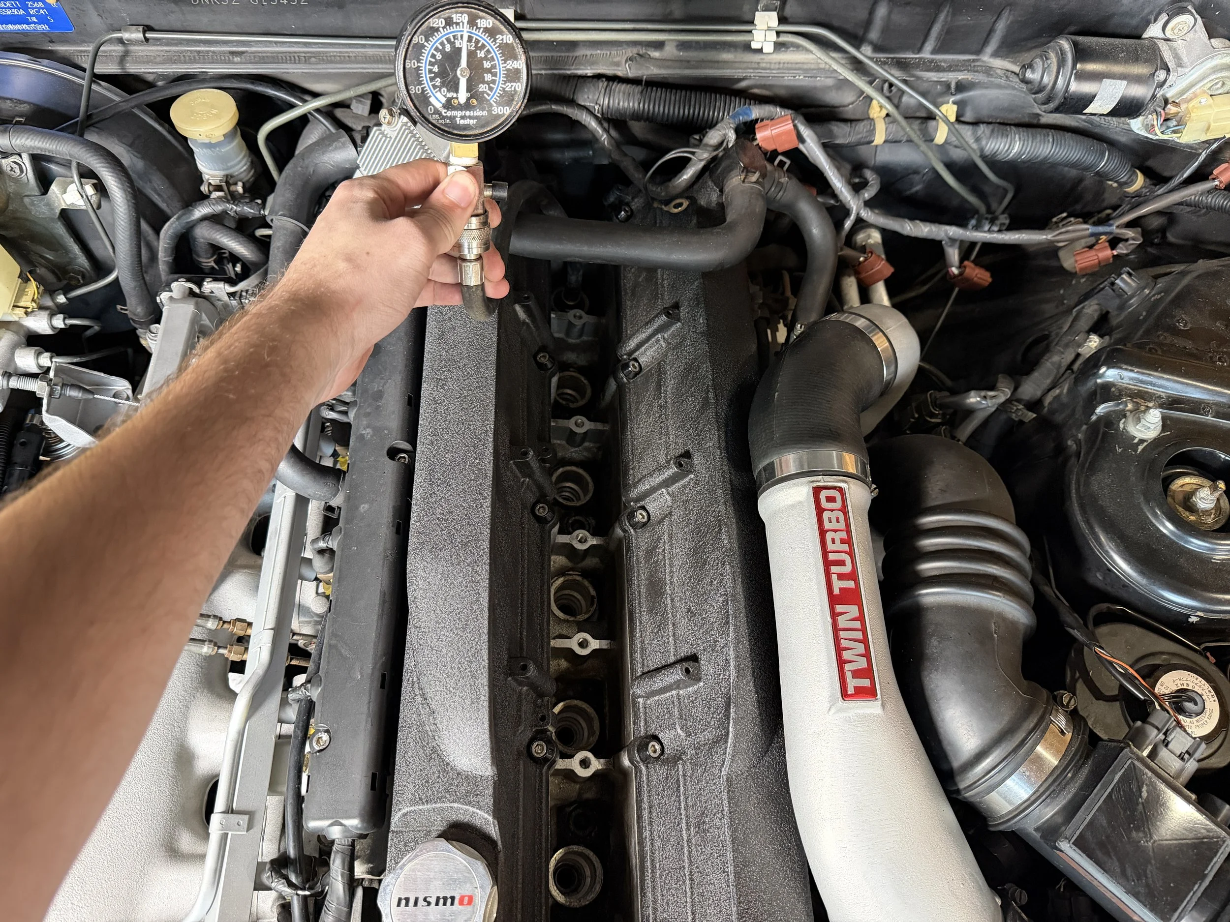 Person testing the turbo pressure with a gauge on a car engine, with visible turbo labeled 'Twin Turbo' and engine components.