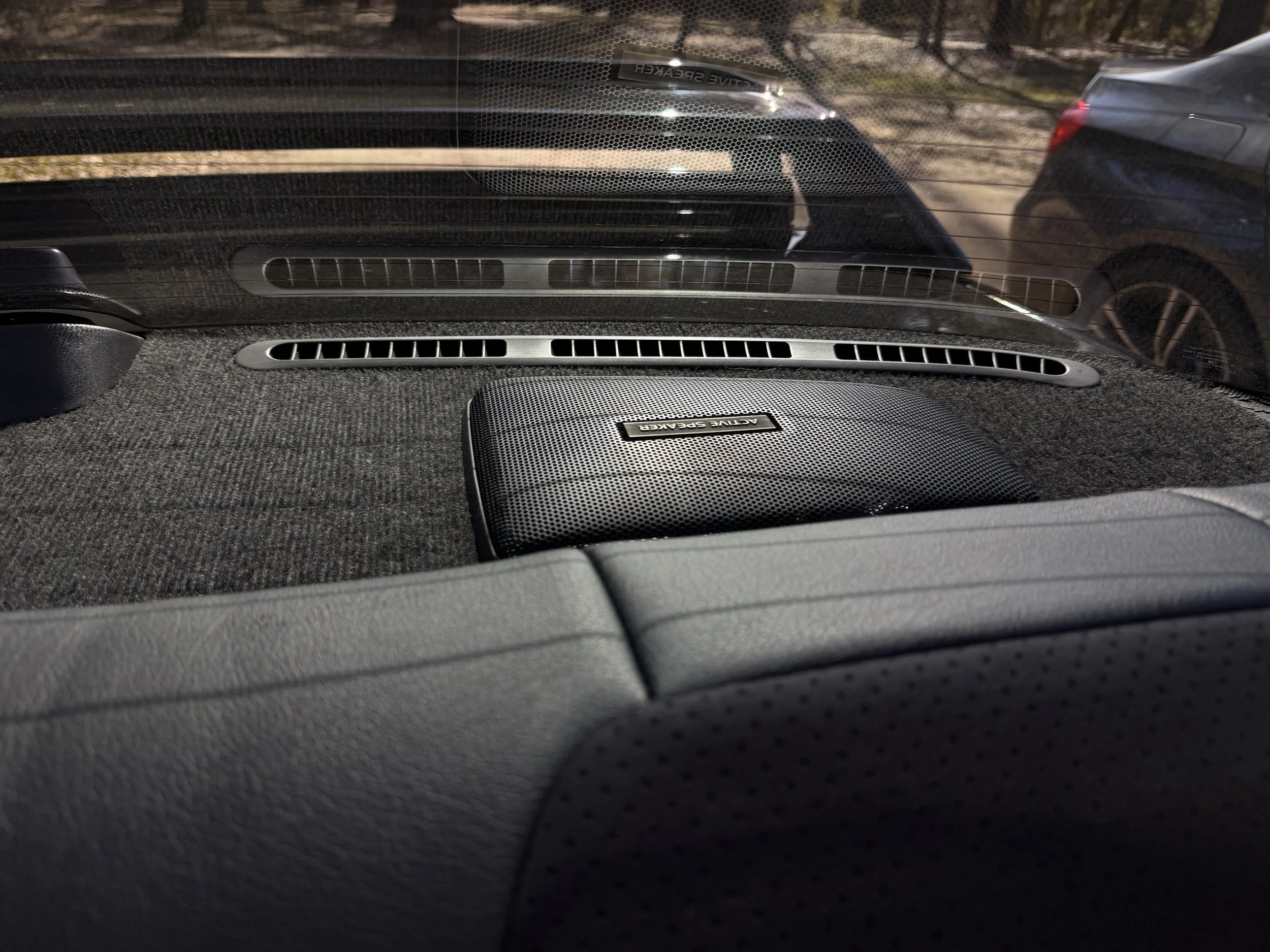 View from inside a vehicle showing a custom audio speaker on the dashboard, with the rear window, dashboard vents, and parked cars visible outside.