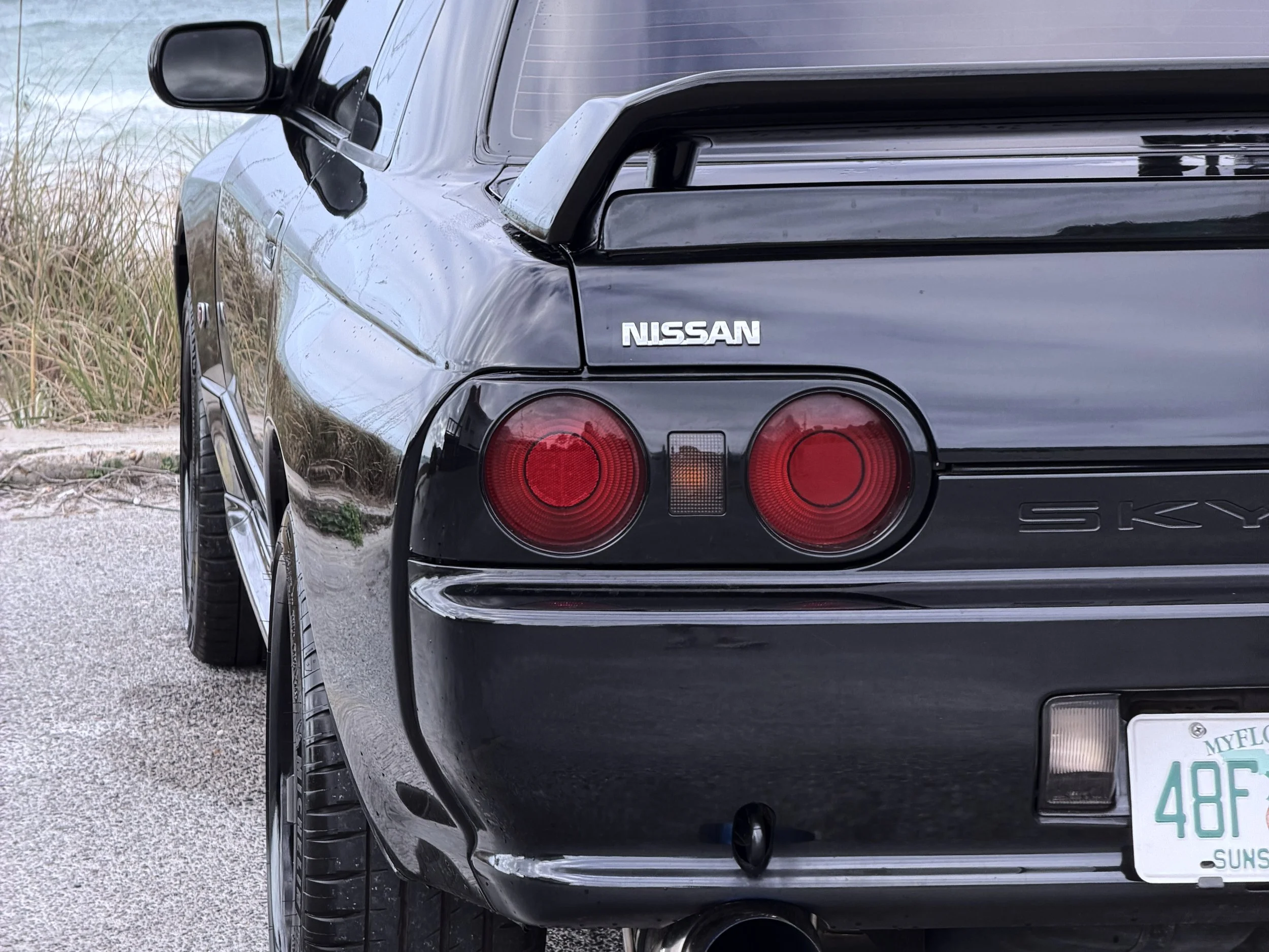 Close-up of the rear end of a black Nissan Skyline car, showing the tail lights, license plate, the Nissan badge, and part of the rear wing, parked on a gravel surface with grass and plants nearby.