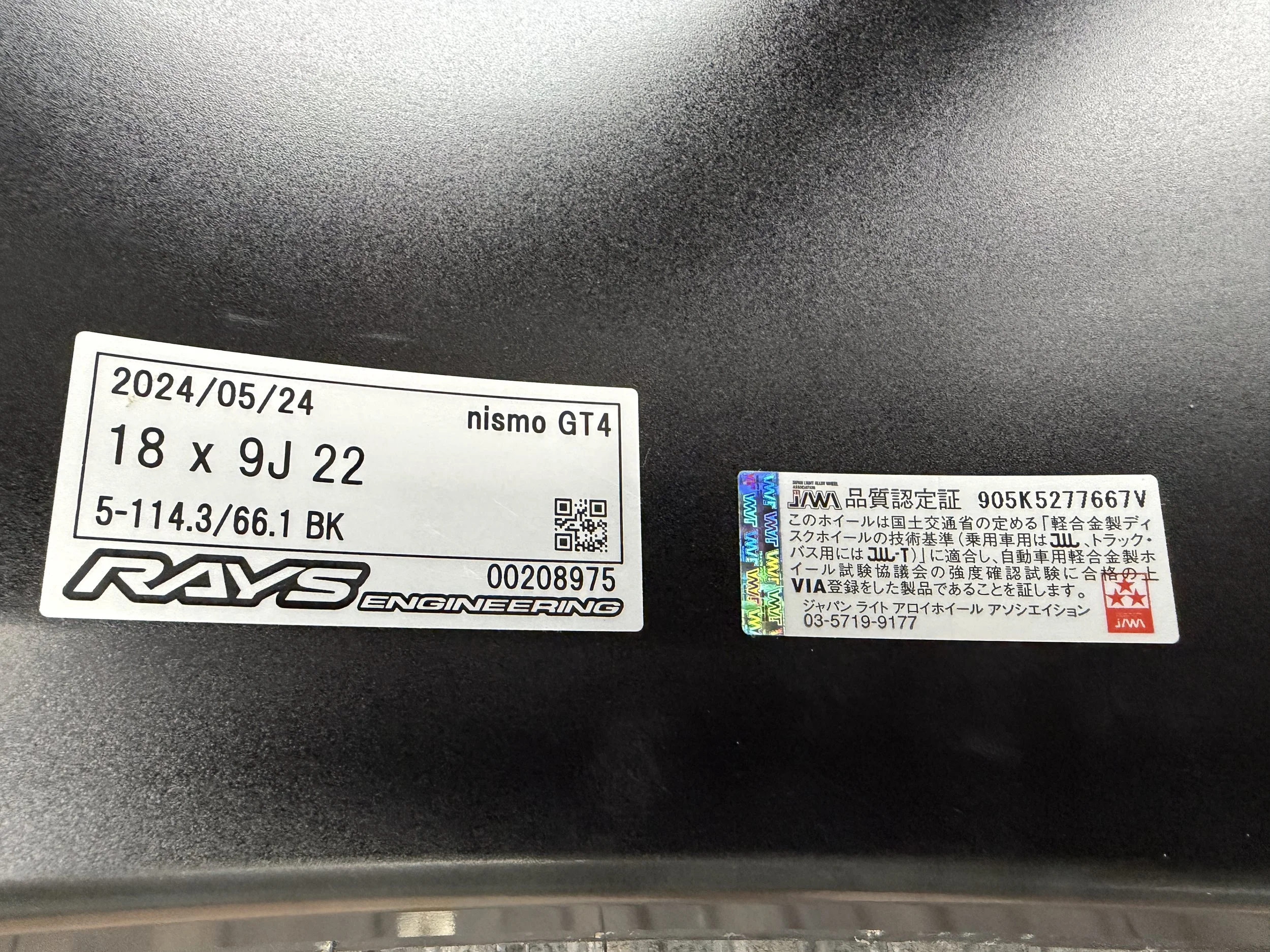 Close-up of a tire with two labels: one from RAYS Engineering indicating the tire size 18x9J22, date 2024/05/24, and another from a Japanese certification authority with Japanese text and a holographic sticker.