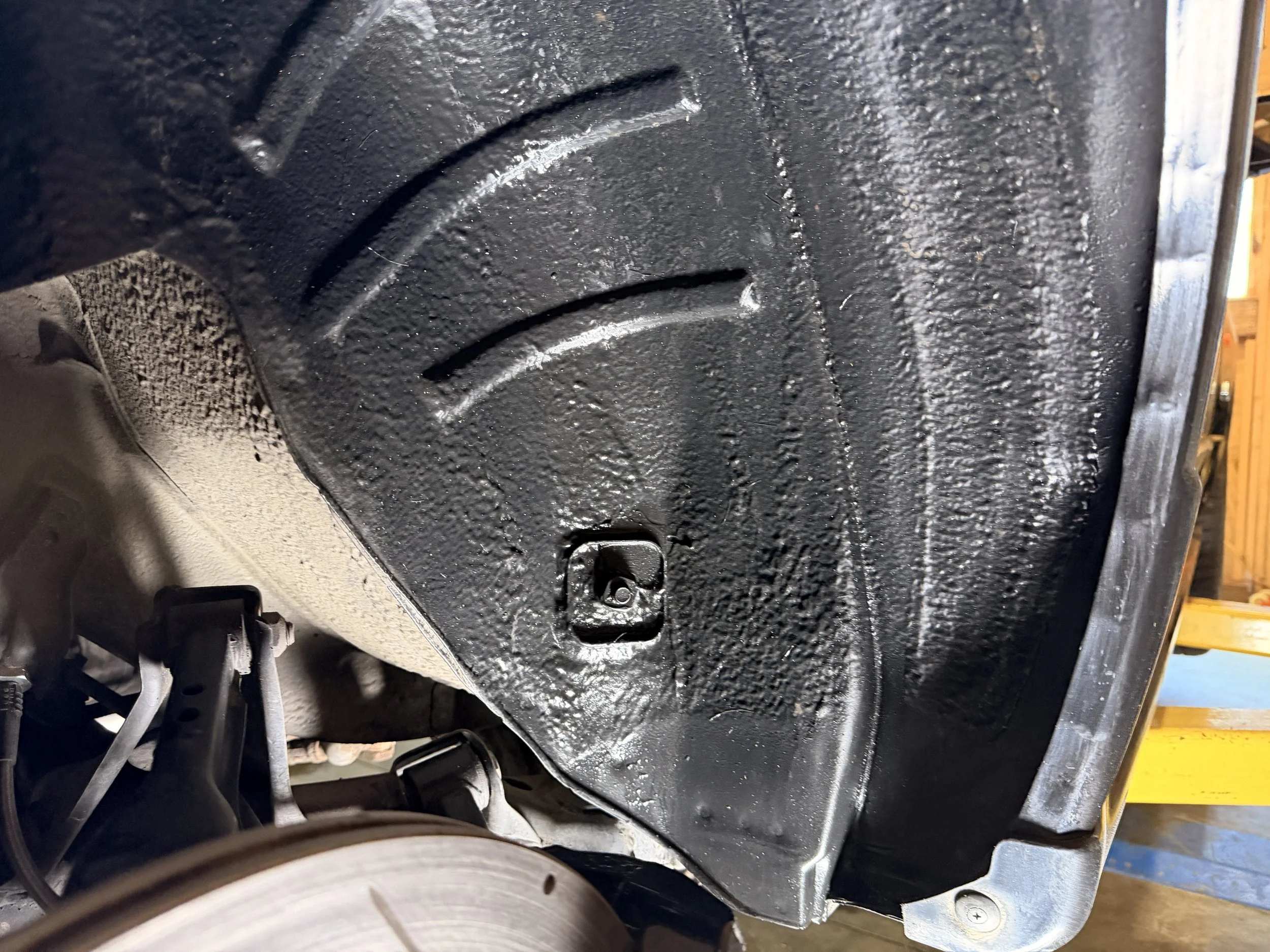 Underbody of a vehicle showing a freshly coated black undercoating, with a part of the wheel and suspension visible on the left side.