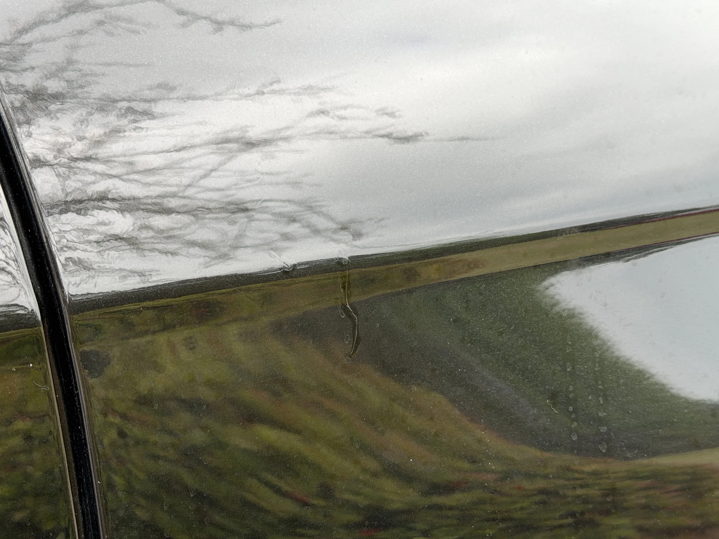 Close-up of a black car door with a small scratch and reflection of a cloudy sky and trees.