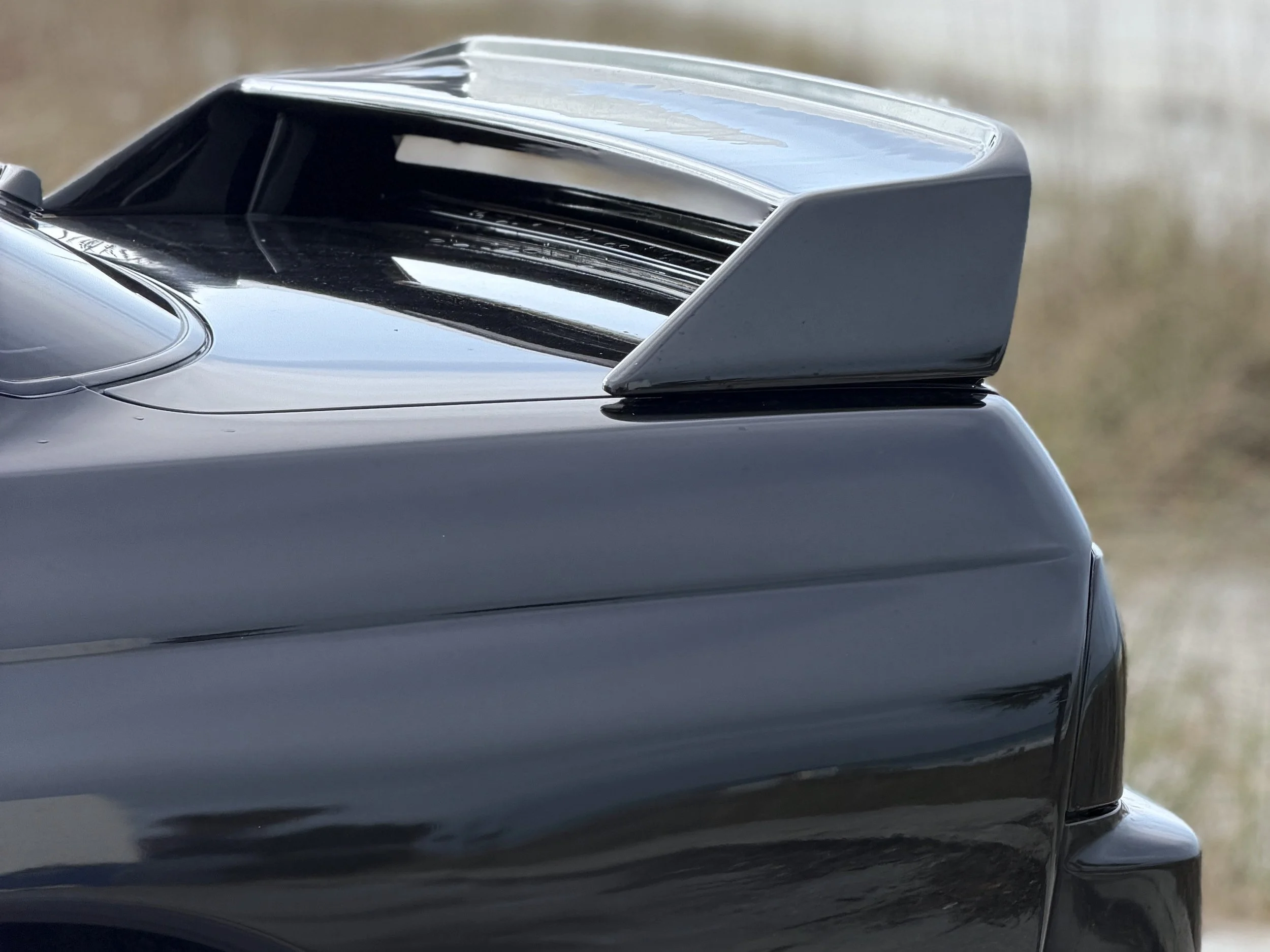 Close-up of a car's rear spoiler and trunk, painted black with a glossy finish, on a blurred outdoor background.