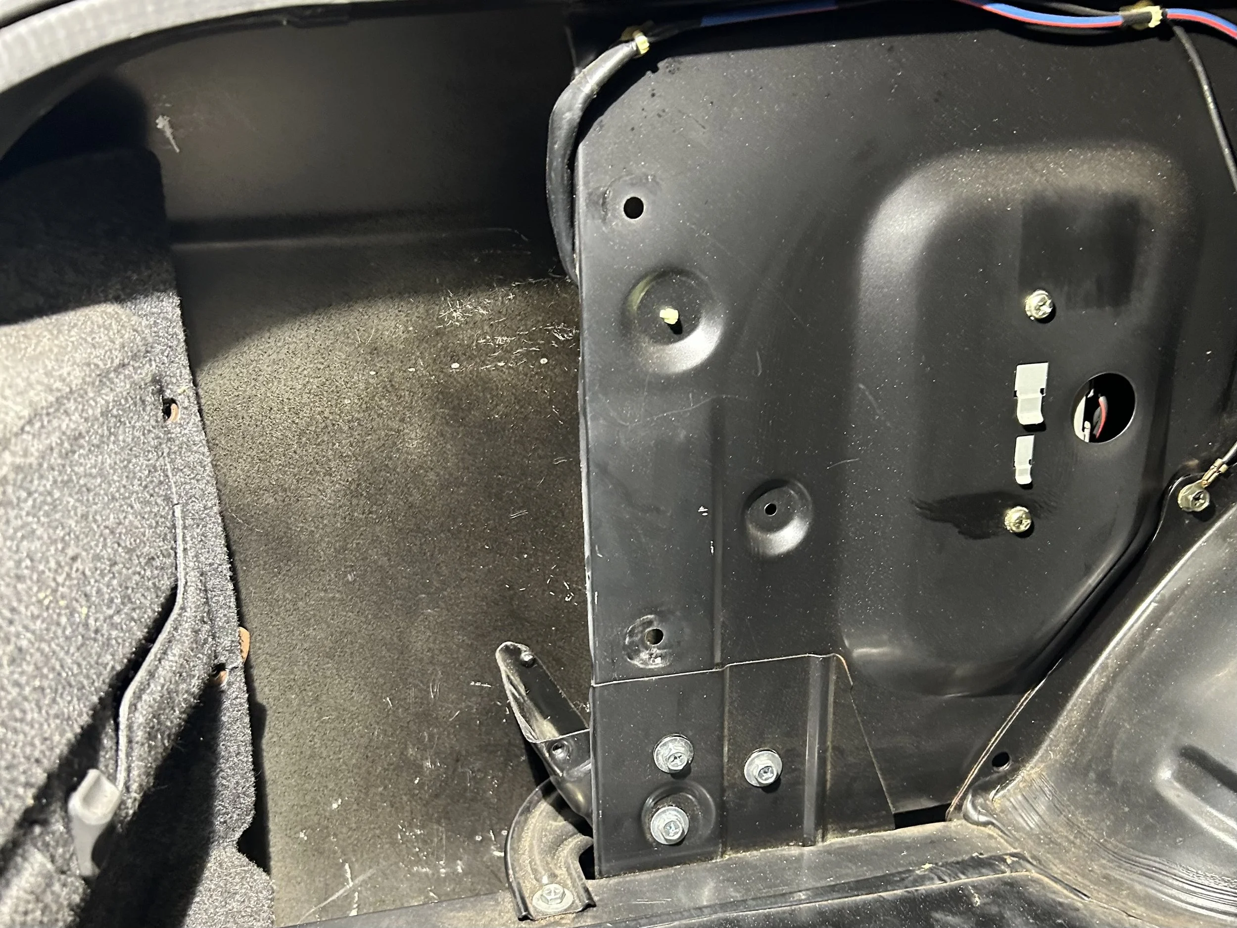 The interior of a vehicle's trunk with black plastic panels removed, exposing the inner structure and wiring.