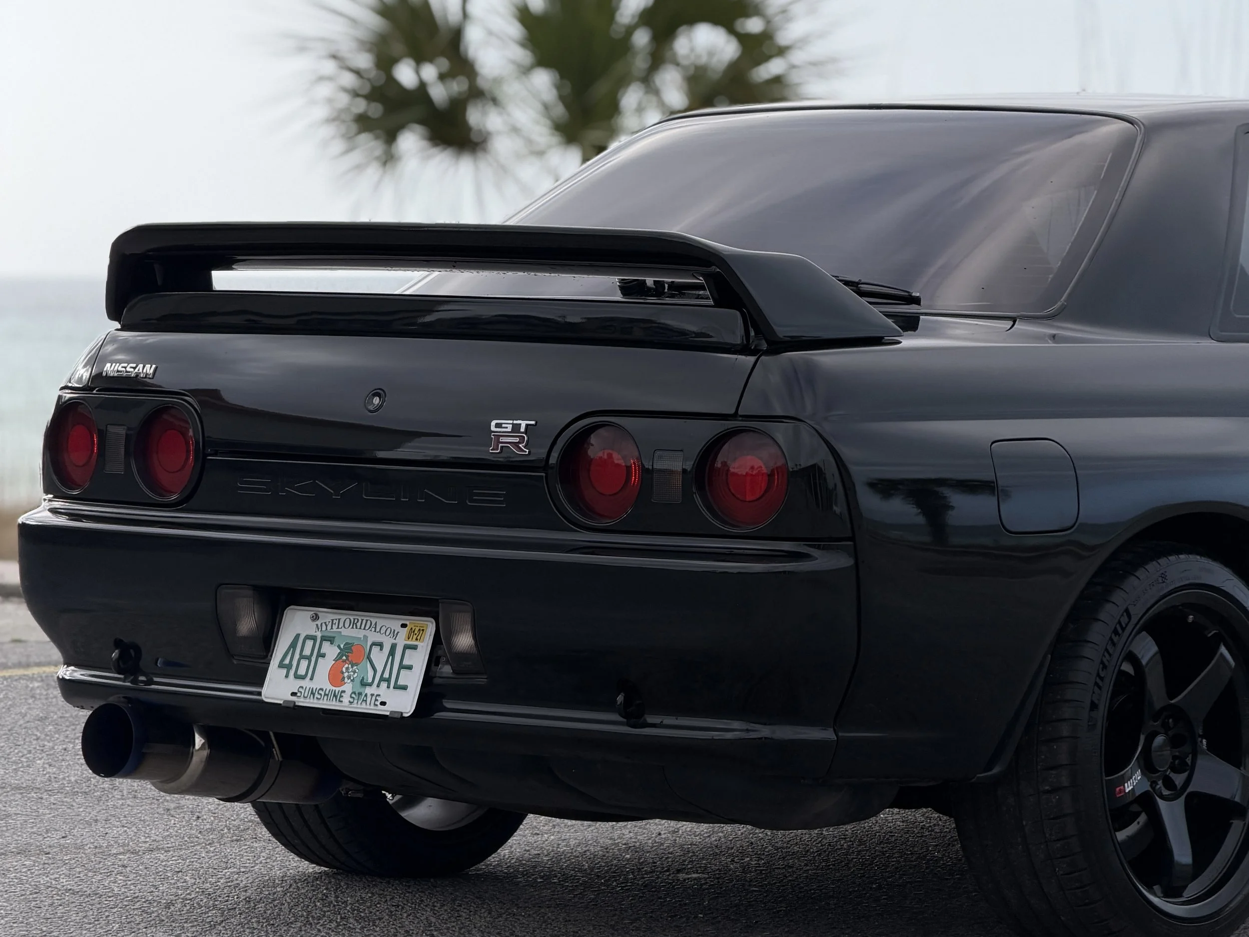 Rear view of a black Nissan Skyline GT-R car with a spoiler, red tail lights, and Florida license plate reading 48F SAFE, parked outdoors.