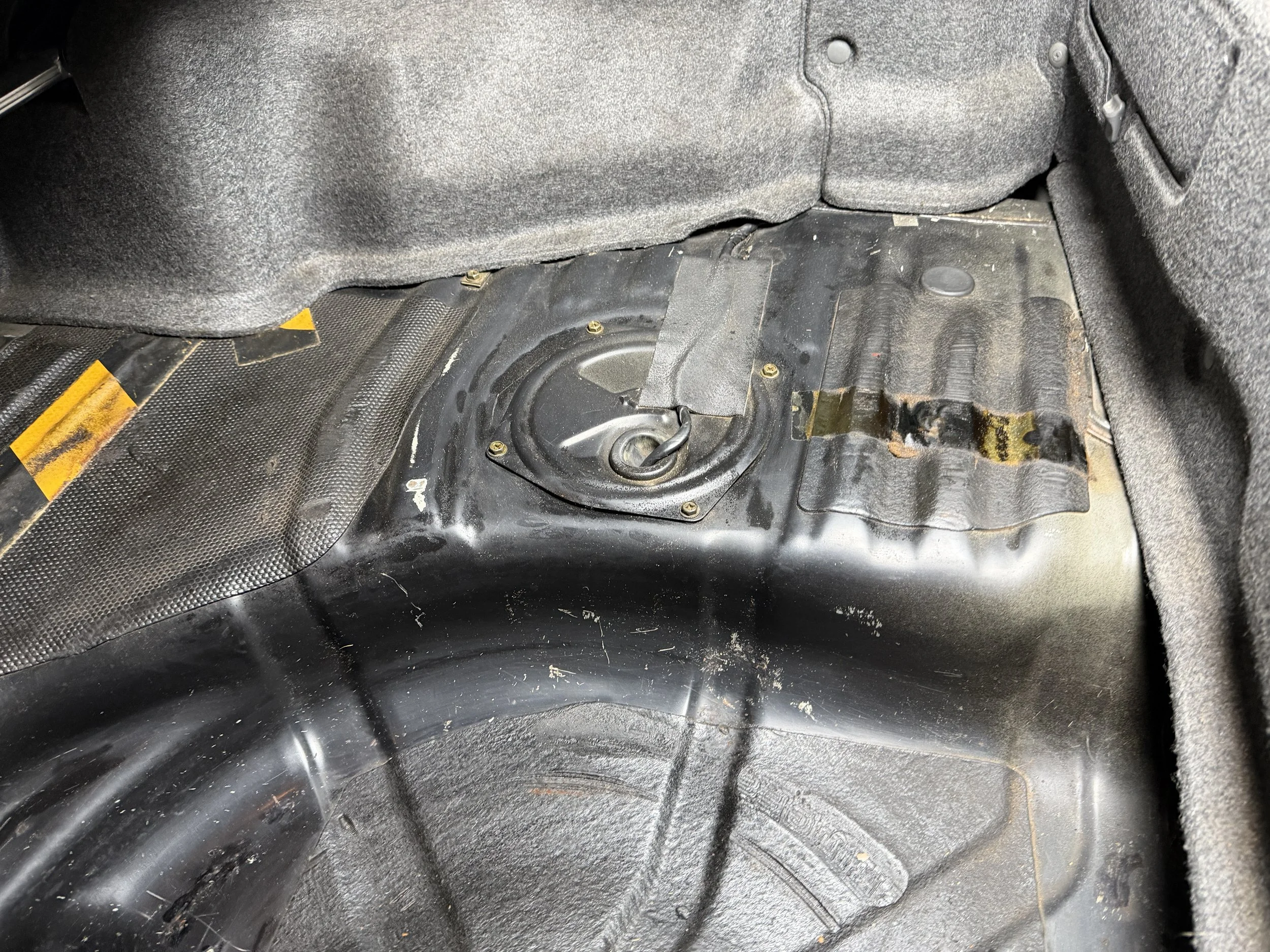 The image shows the inside of a car trunk with the floor cover removed, exposing the spare tire well and a circular mounting point.