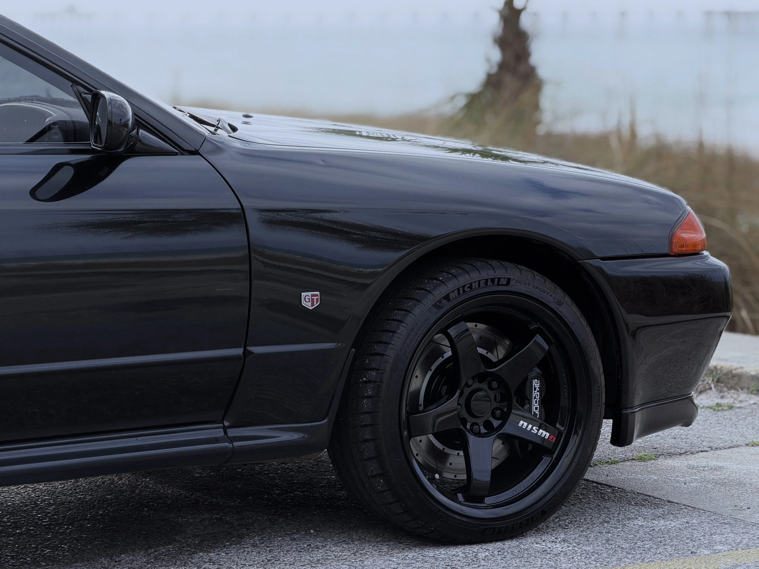 Close-up of the front side of a black sports car with a GT badge and racing wheels, parked on a gravel surface near a body of water with a tree in the background.
