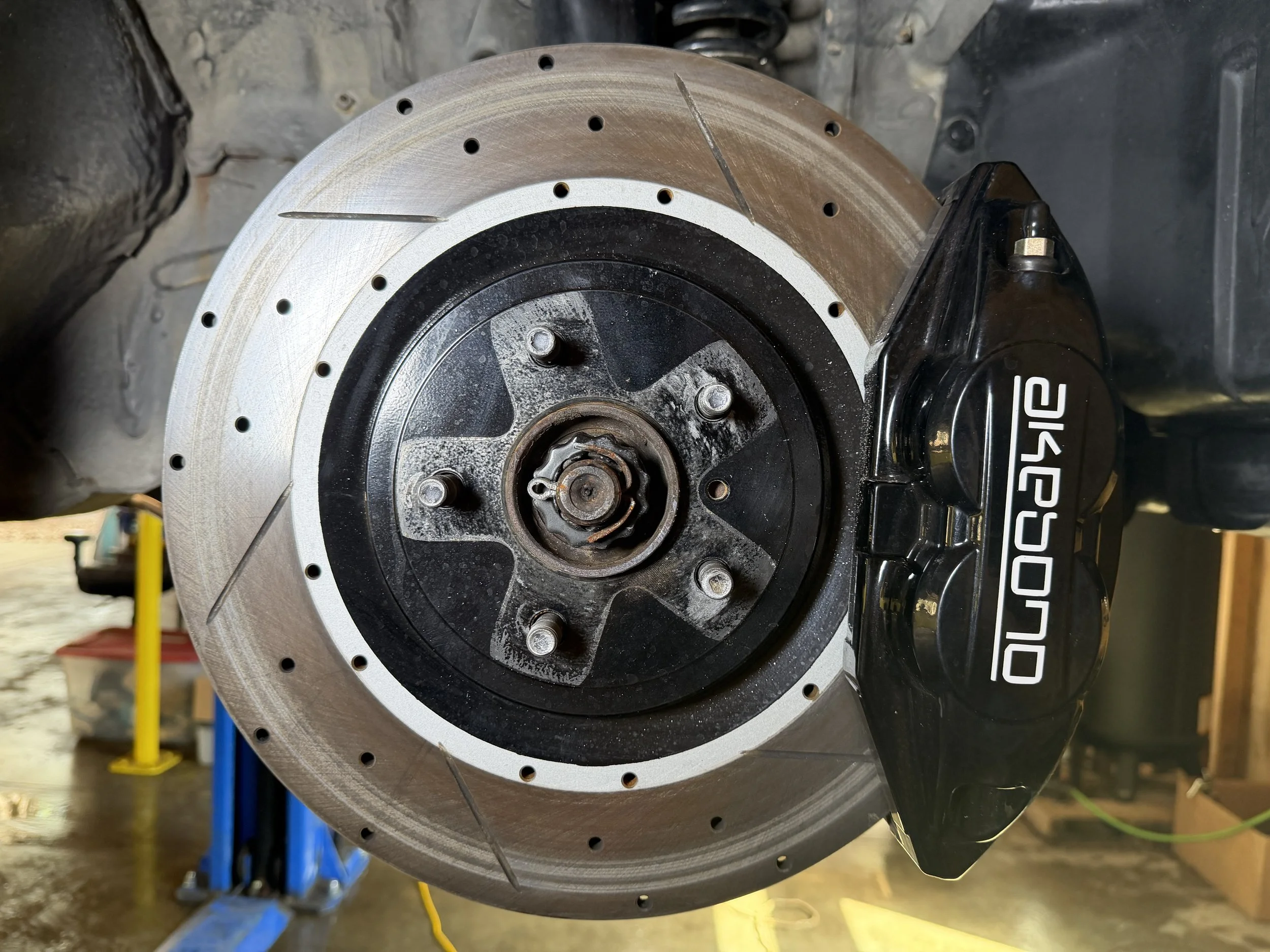 Close-up of a car brake rotor and caliper, with the brand name 'Dakota' on the caliper, in an automotive workshop.