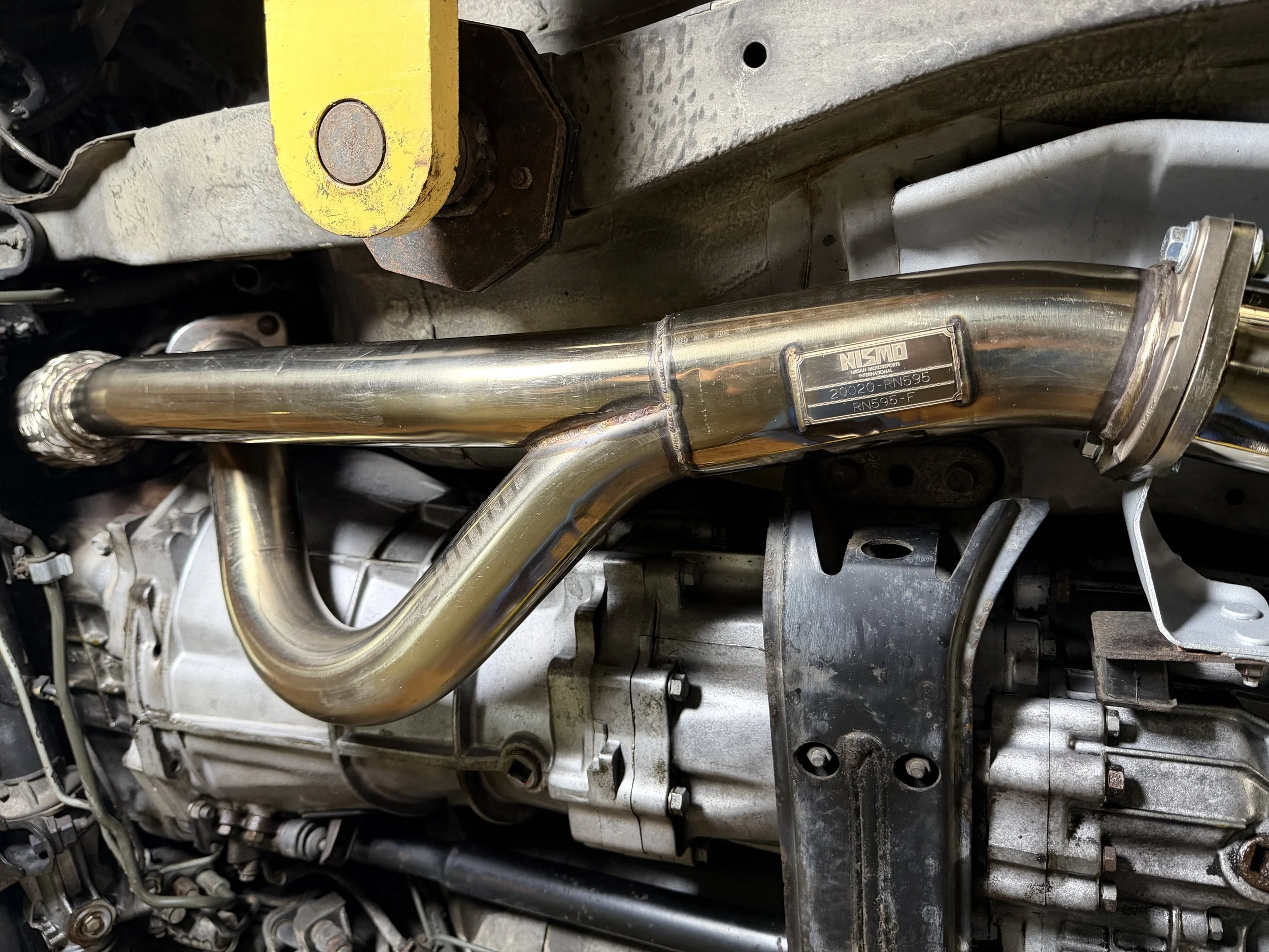 Close-up of a metallic pipe or tube connected to a car engine, with a label reading NISMO, 20020-RN595, and RN595-F.