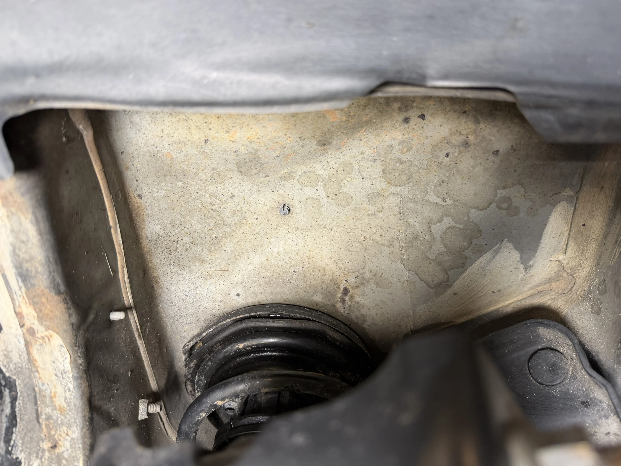 Inside view of a vehicle's engine compartment showing a coil spring and metal surface with dirt and oil stains.