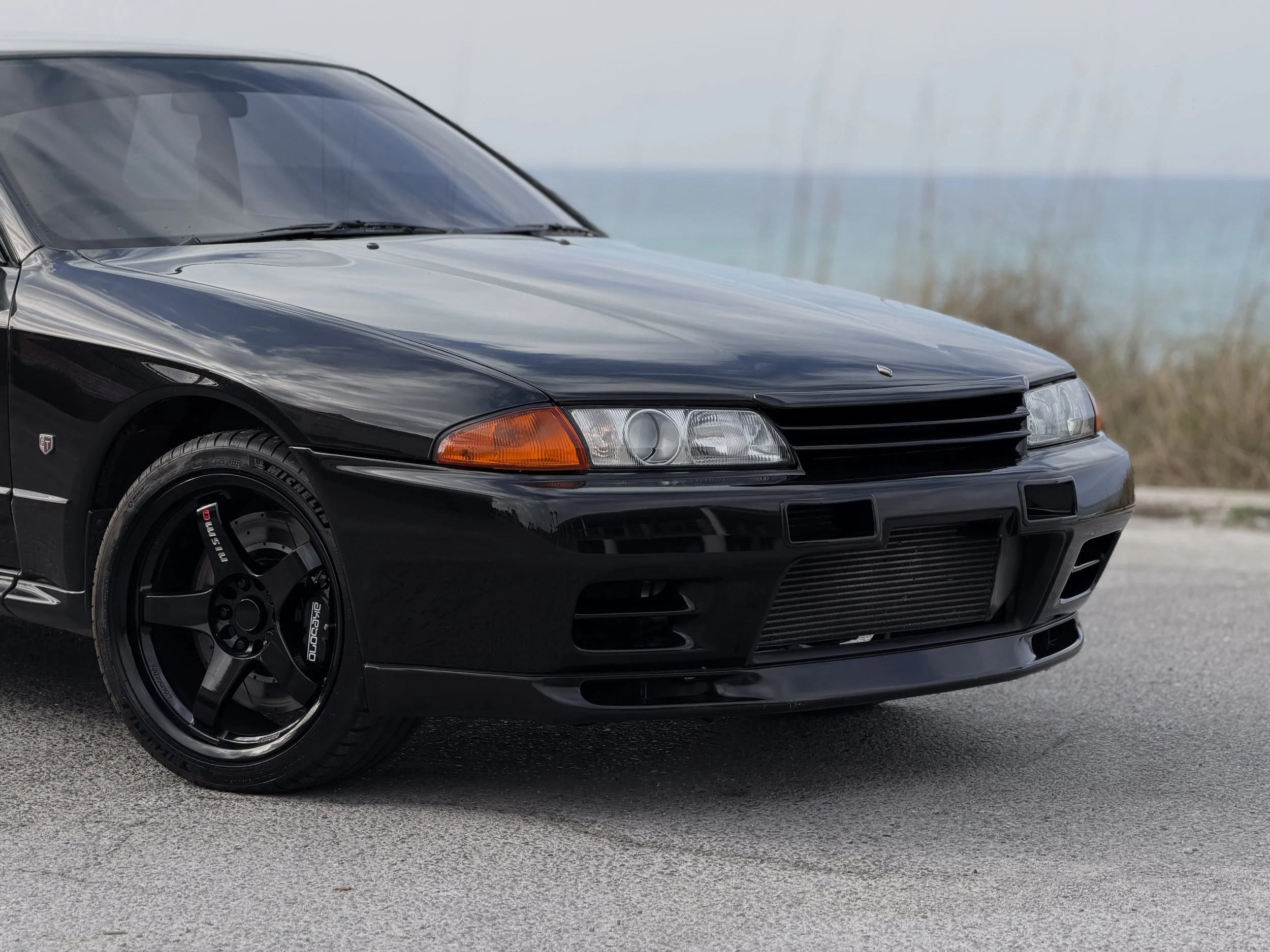 R32 NISSAN SKYLINE GTR FOR SALE
Black sports car parked on a paved road near the coast with grass and the ocean in the background.