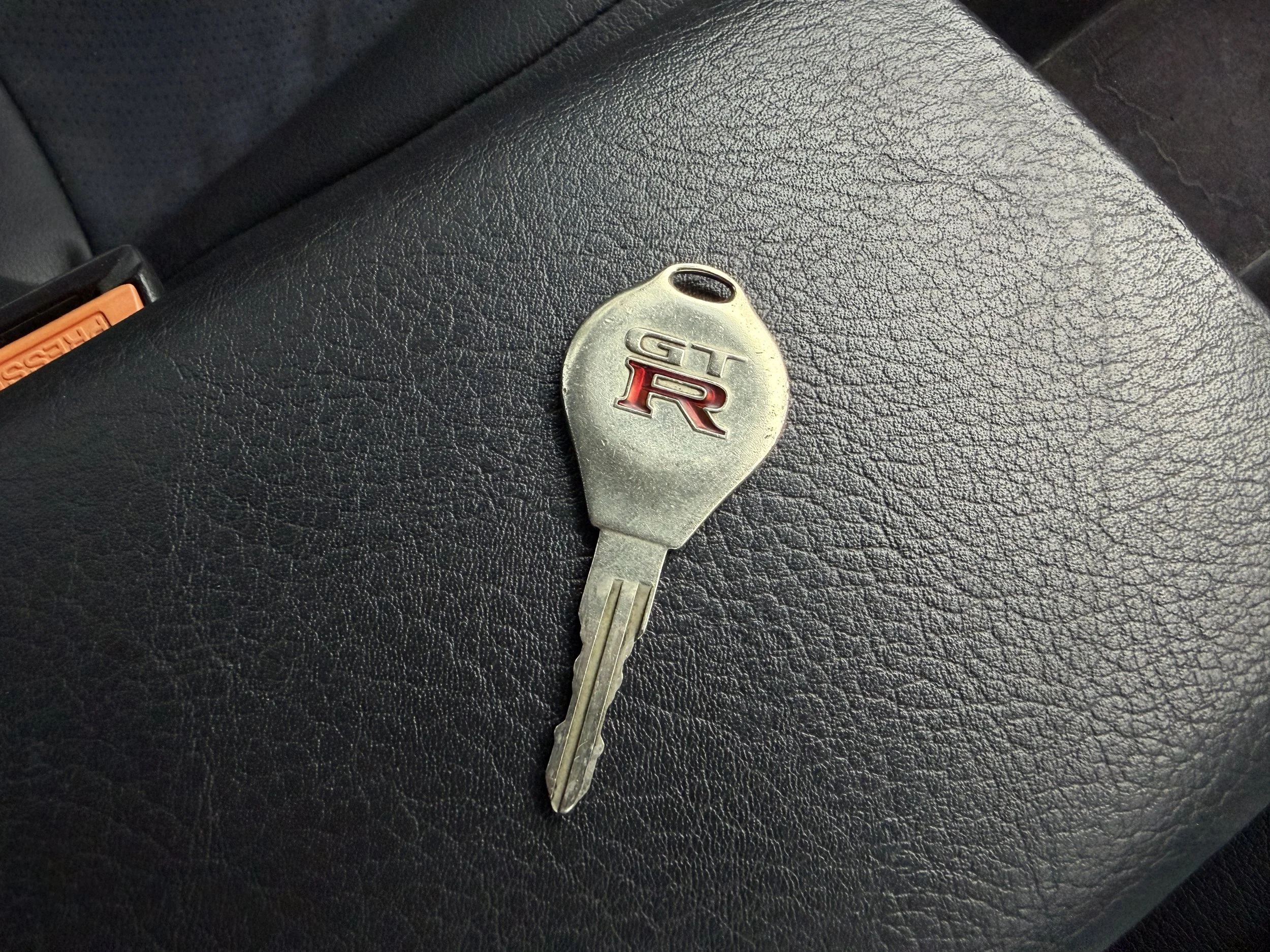 A car key with a metal head, engraved with the words 'GT' and 'R' in red and black, resting on a textured black surface inside a car.
