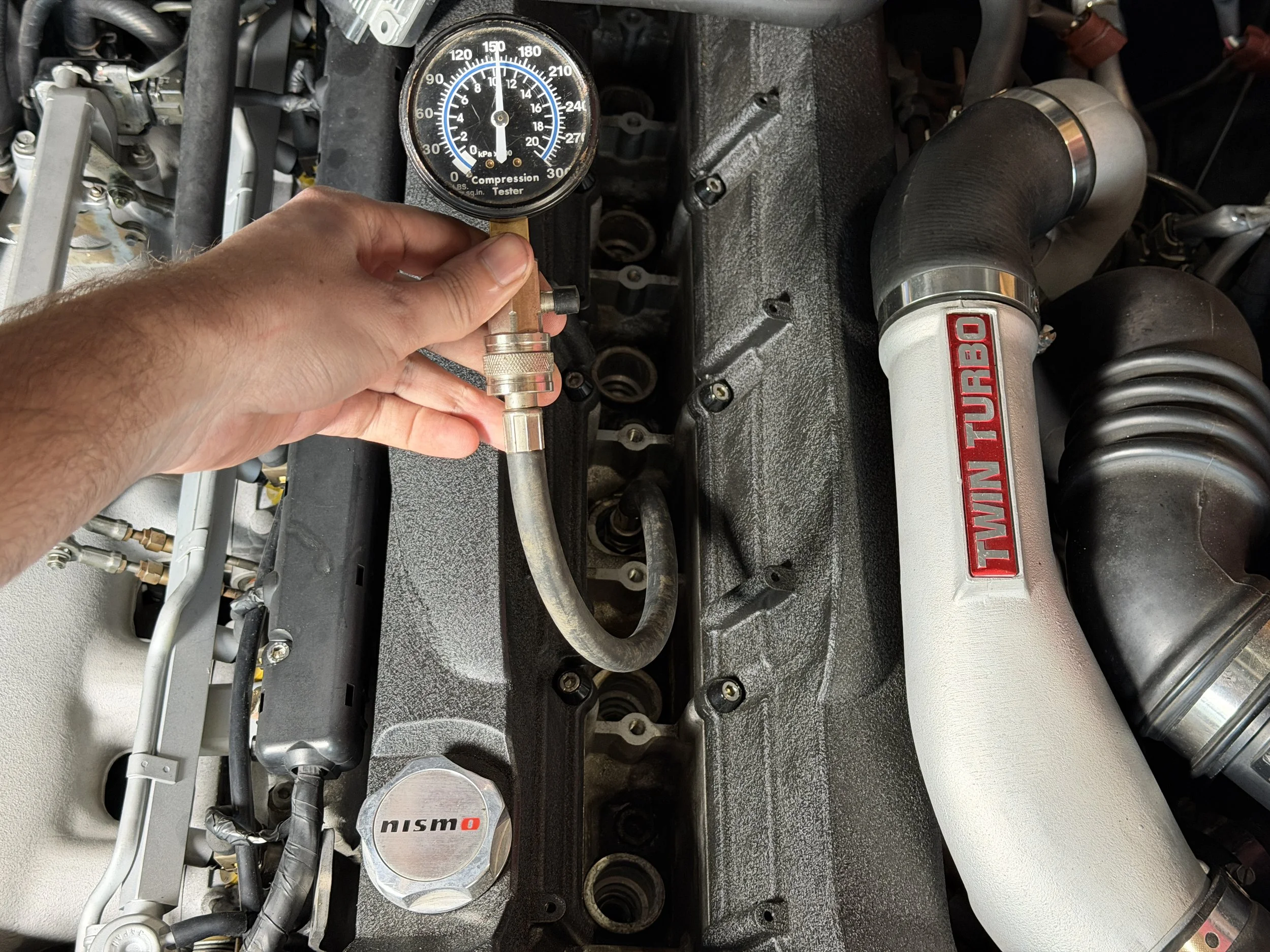 A person is measuring the pressure in a car's engine with a gauge. The engine has a twin-turbo setup, with visible branded components including a Nismo valve cover and a large white intercooler pipe.