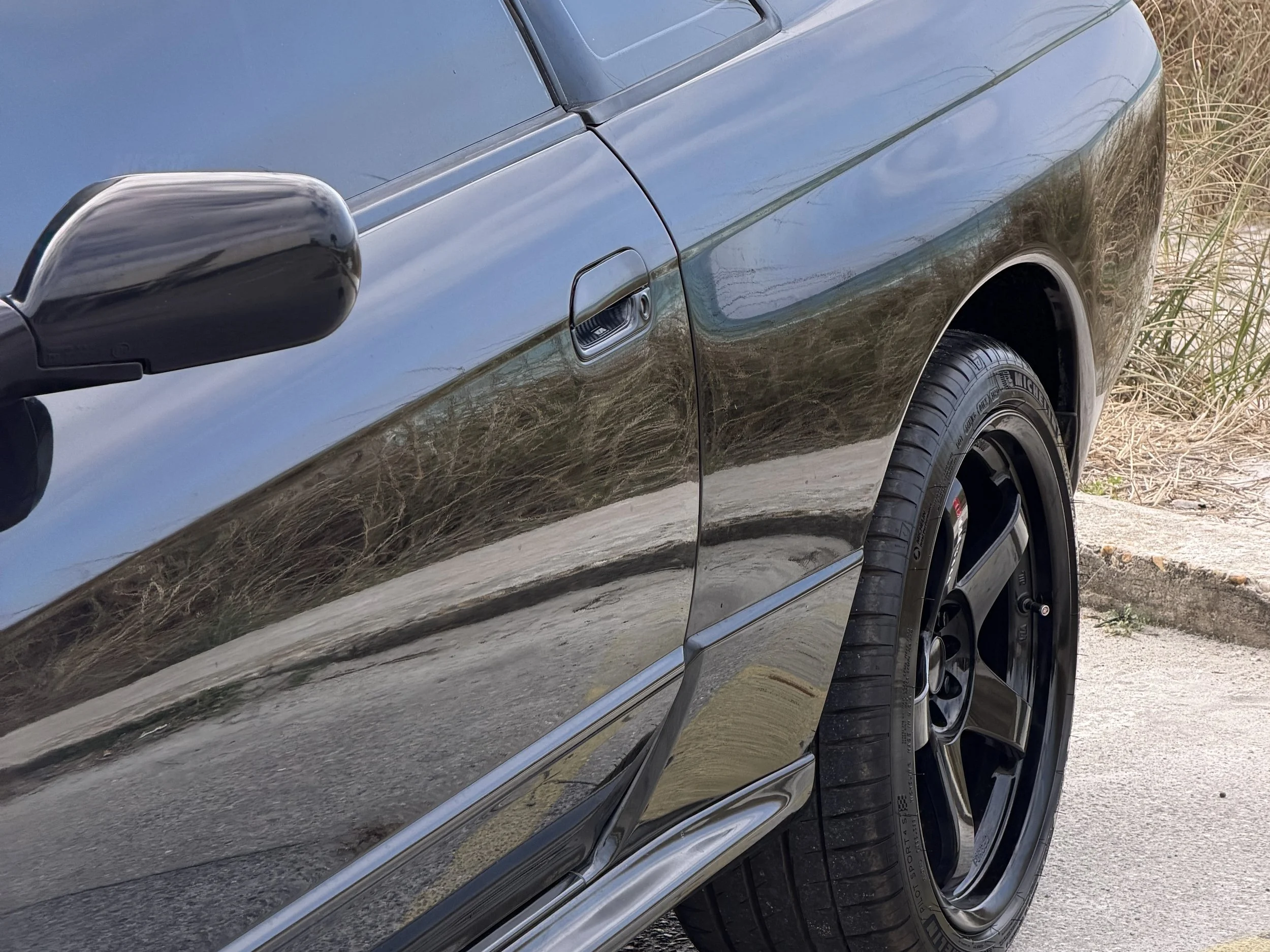 Close-up of a black car's side showing the door, side mirror, and front wheel on a dirt and grass surface.