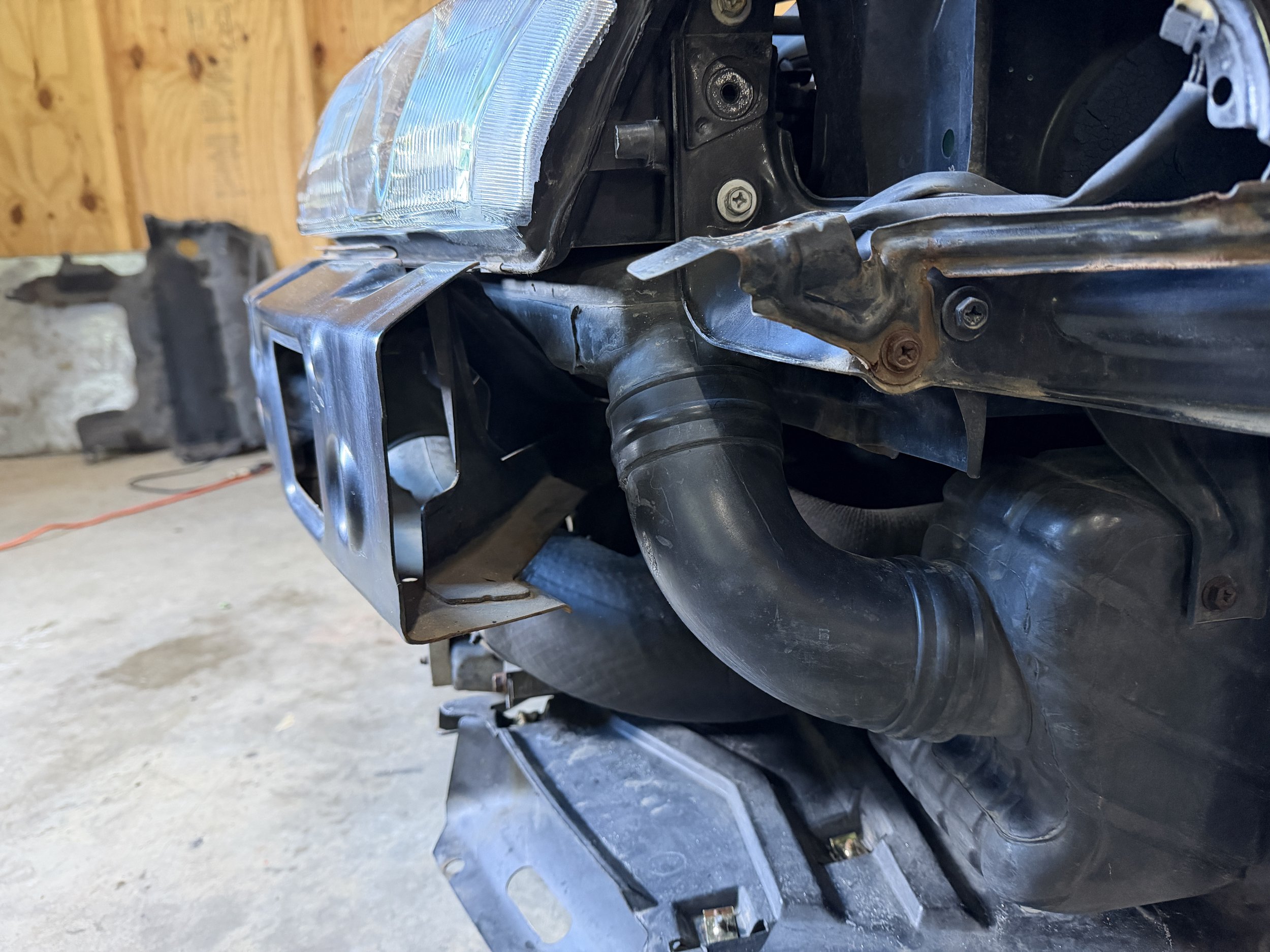 Close-up of a car front end with a headlight, metal mounting brackets, a black plastic air intake hose, and part of the engine bay in a garage with a wooden wall.