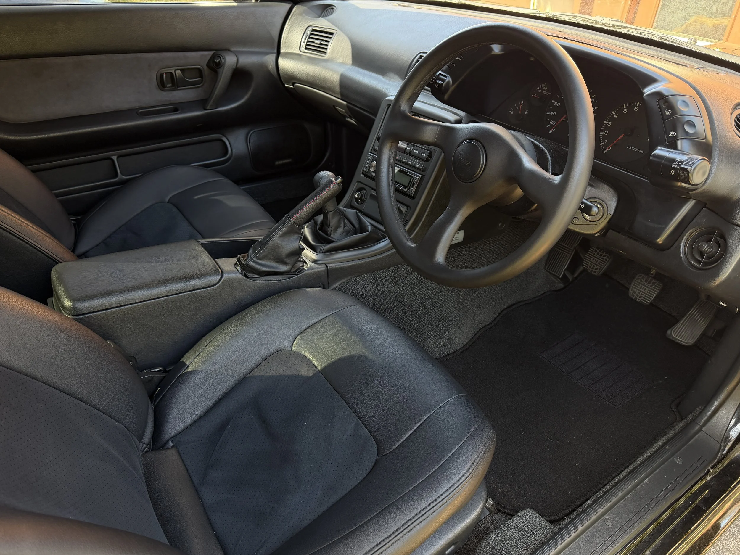 Interior of a car showcasing black leather seats, a manual gear shift, steering wheel, dashboard with gauges, and floor mats.