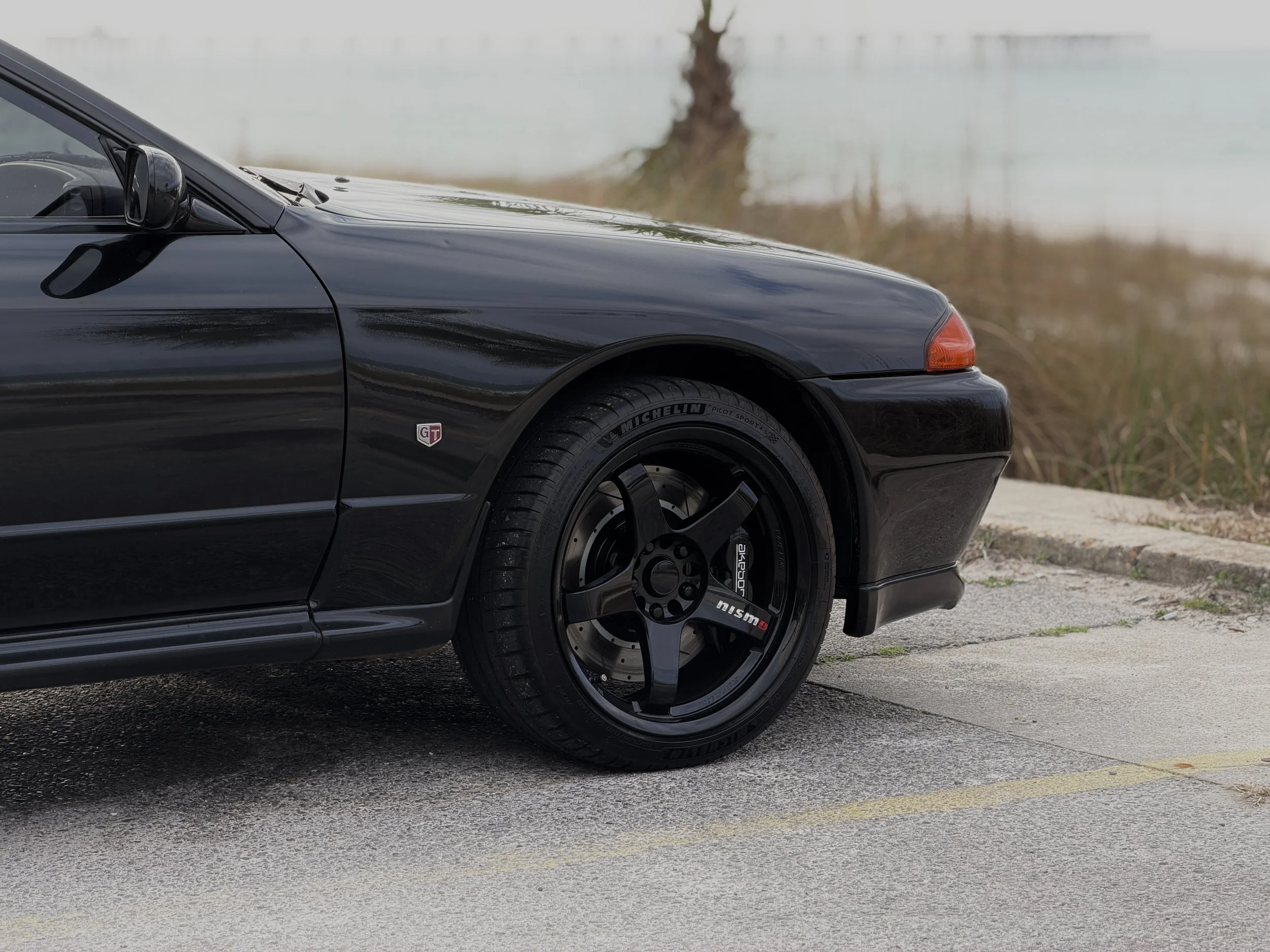 R32 NISSAN SKYLINE GTR FOR SALE
Front part of a black sports car parked on a paved surface with a lake and trees in the background.
