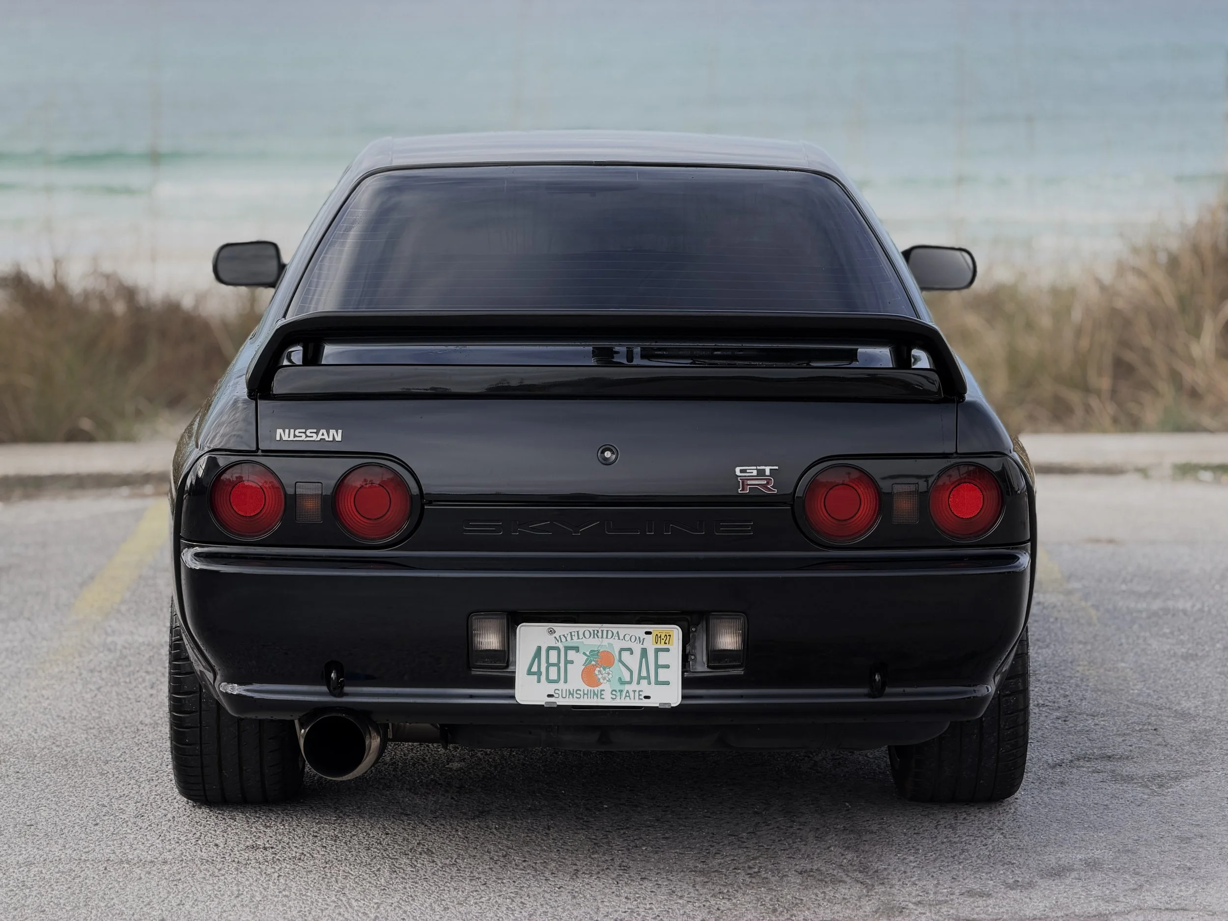 R32 NISSAN SKYLINE GTR FOR SALE
Rear view of a black Nissan Skylines GT-R sports car parked on a paved lot with a beach and ocean in the background.