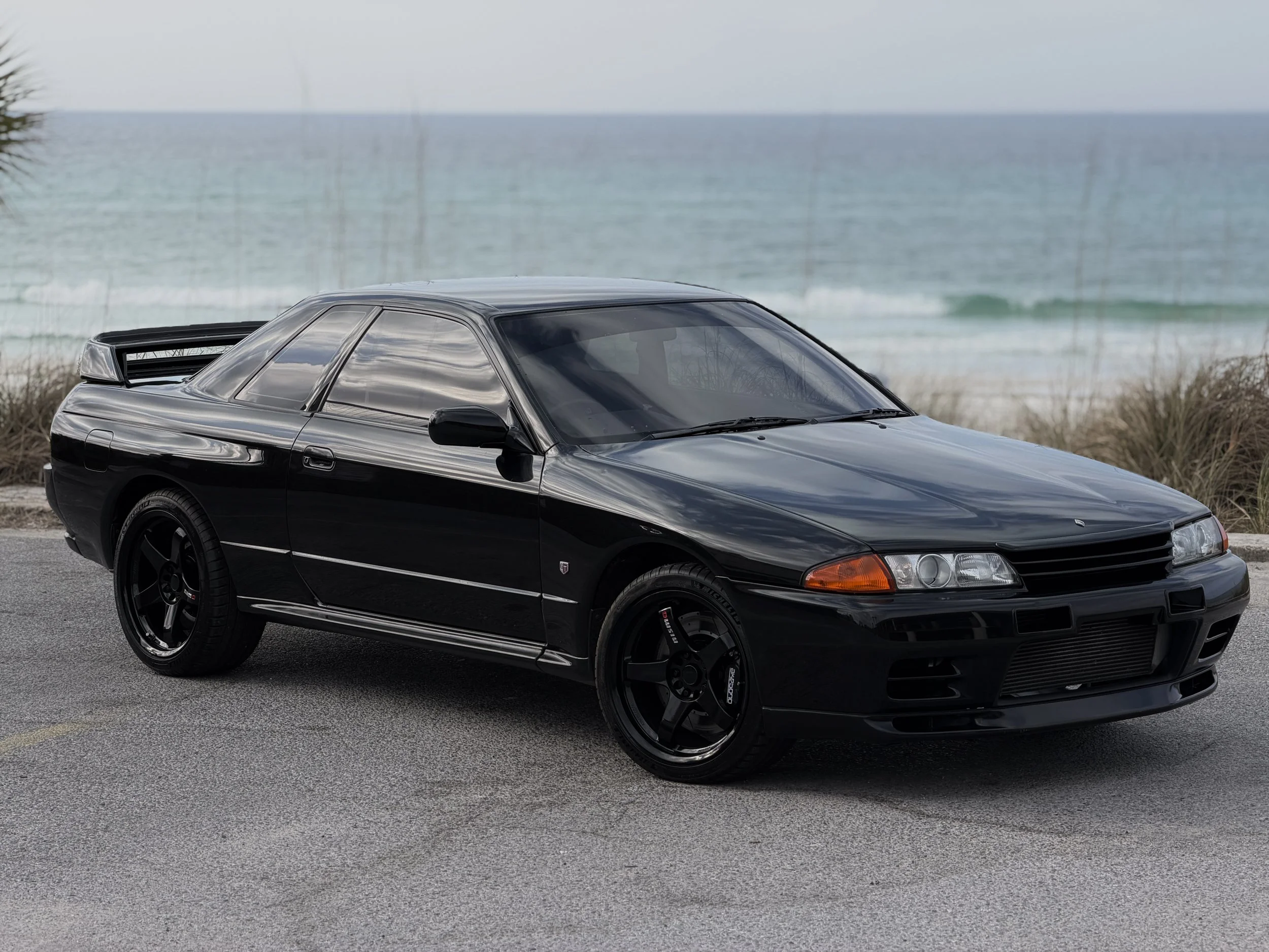 R32 NISSAN SKYLINE GTR FOR SALE
Black classic Nissan Silvia S13 car parked on a road near the beach with ocean waves in the background.