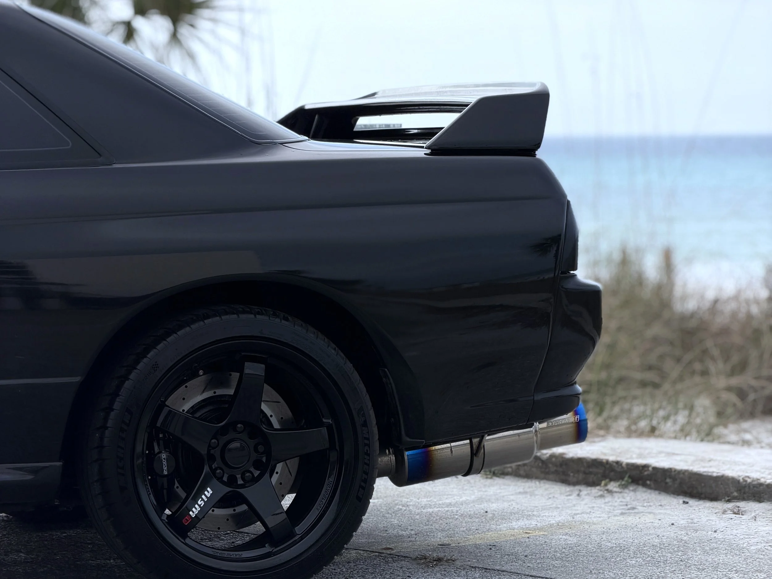 Close-up of the rear side of a black sports car showing the rear wheel and part of the rear wing/spoiler, with a snowy ground and blurred background.