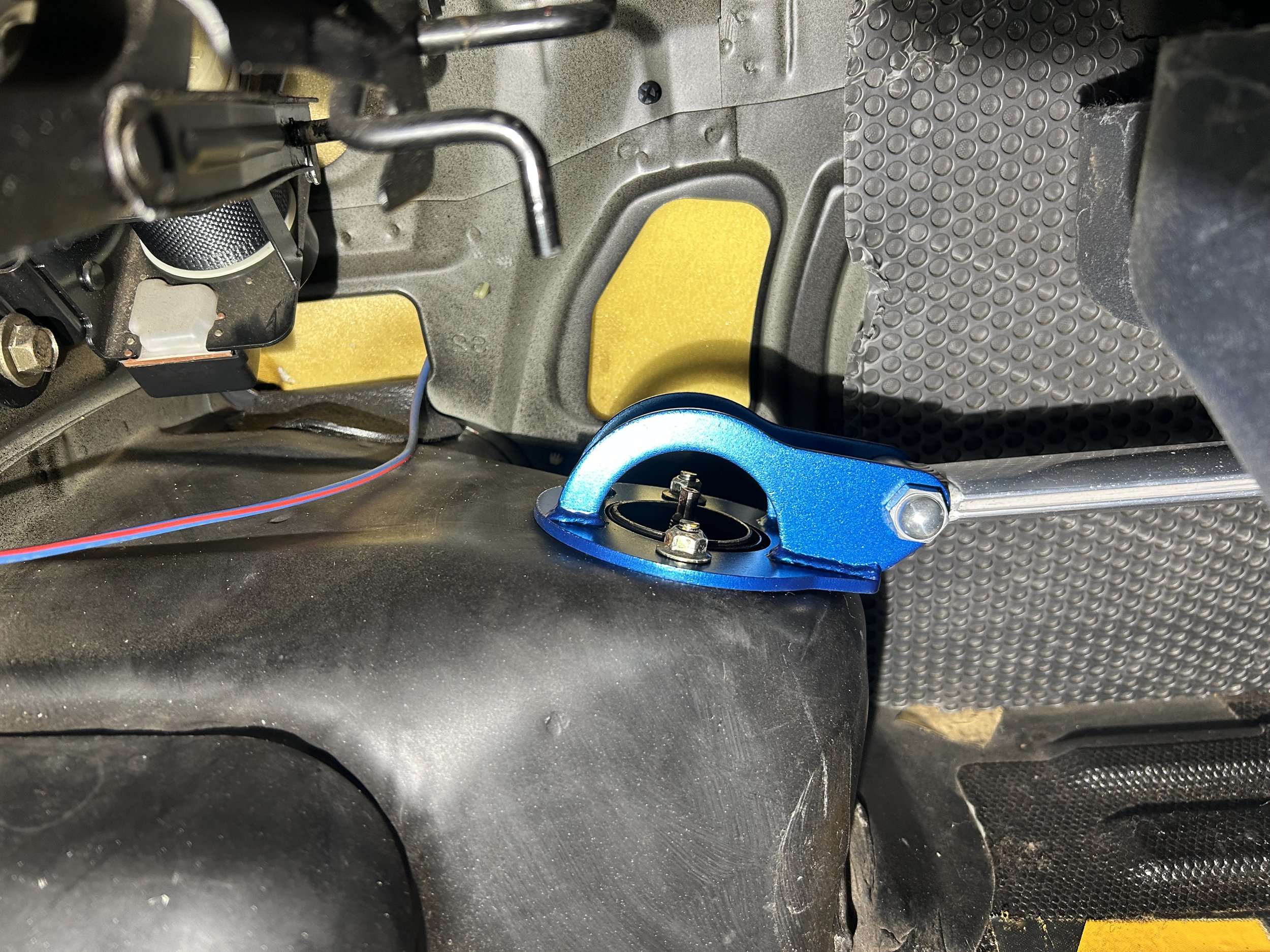 Close-up of a blue metal tool attached to a car's interior, possibly for maintenance or repair, with a black rubber surface and various wires and components around.
