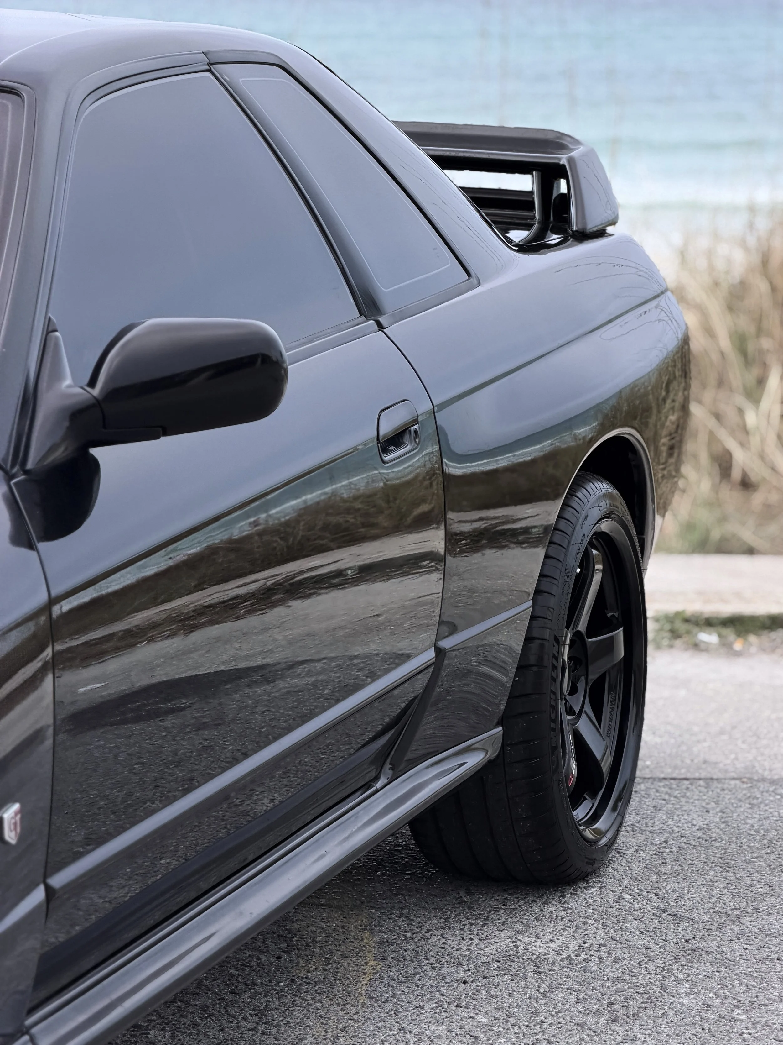 Close-up of the side of a gray sports car with black wheels, parked on a paved surface near a body of water and some grass.