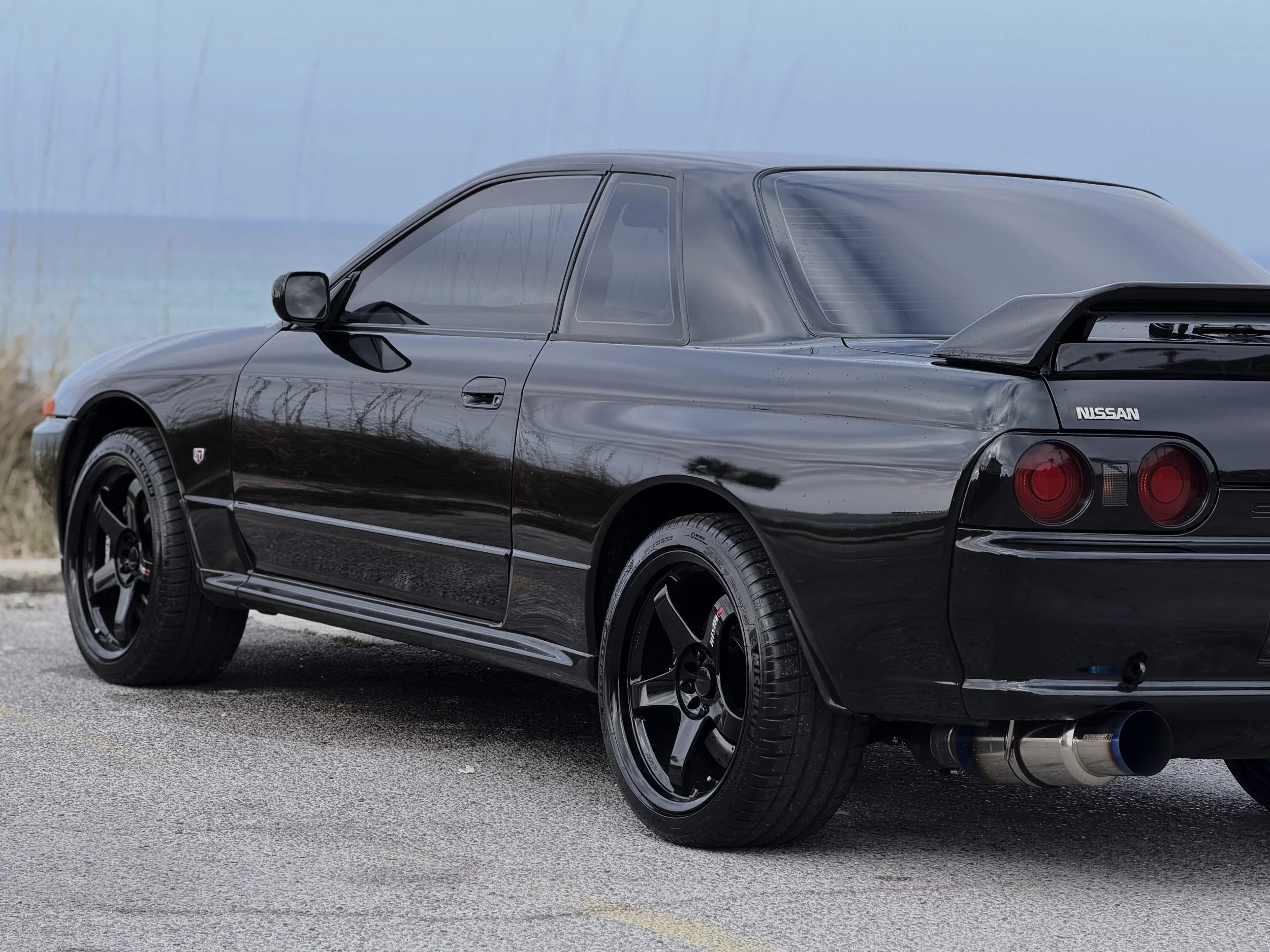 A black Nissan sports car parked on a road with a water body and hills in the background.