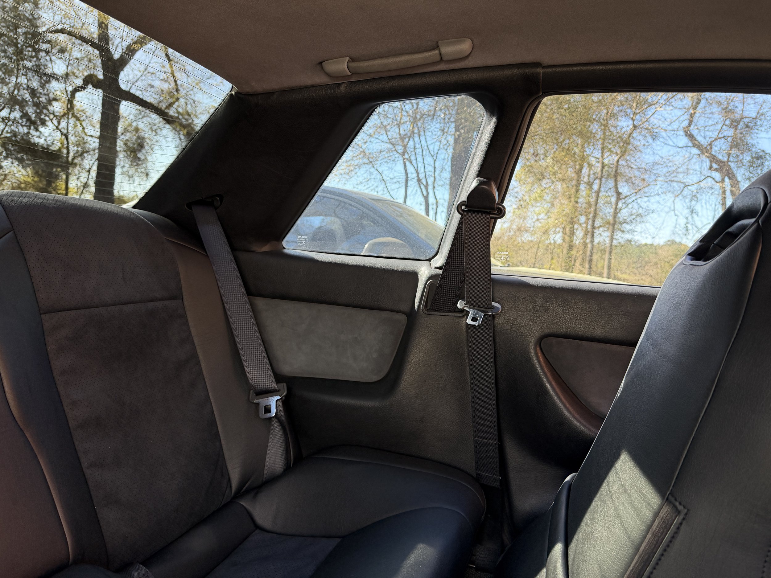Backseat of a vehicle showing two seats with brown and black upholstery, seat belts, and a window with trees visible outside.