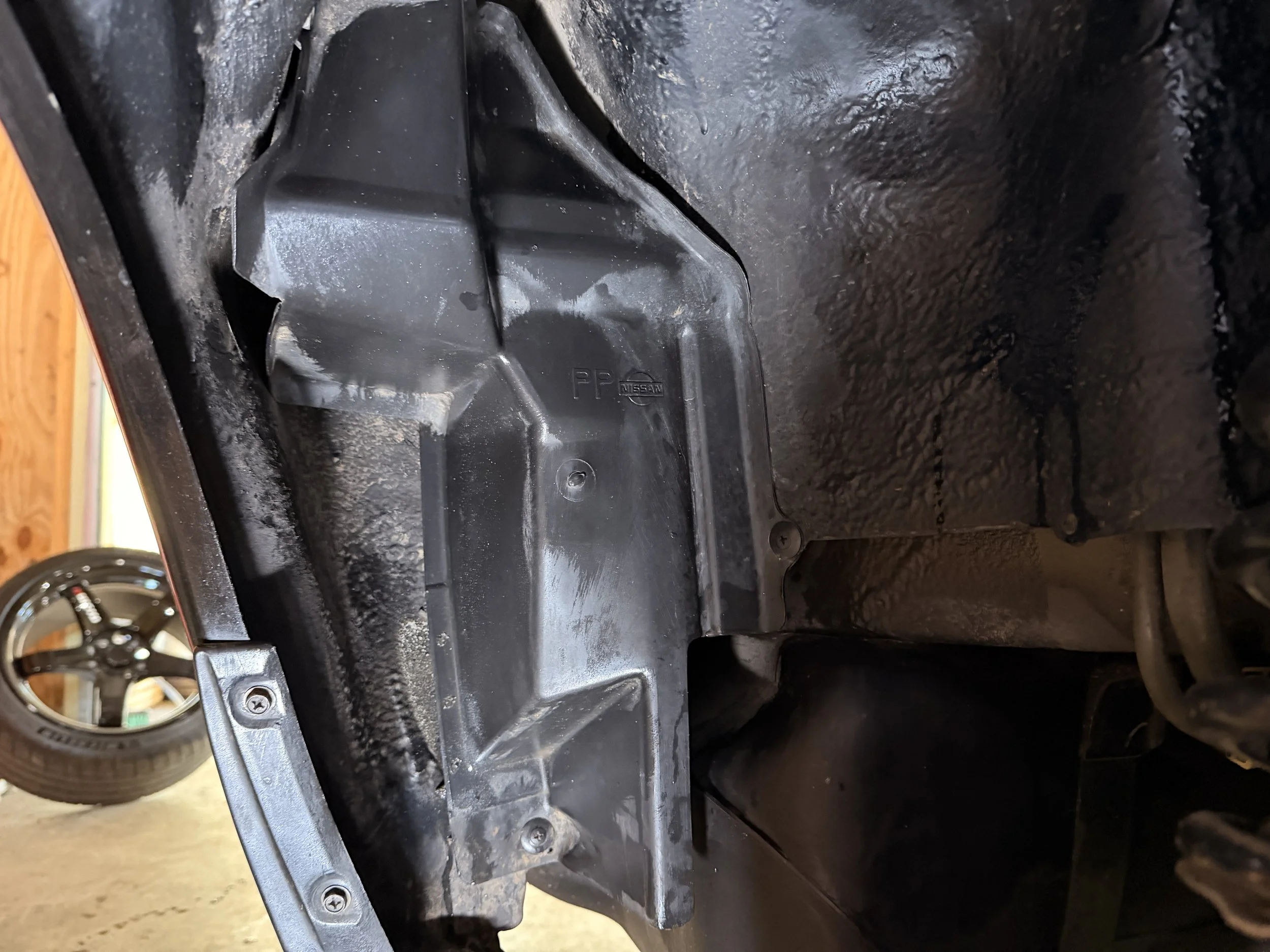 Close-up of a black automotive plastic component with screws, inside a vehicle's undercarriage, with a rear wheel visible in the background.