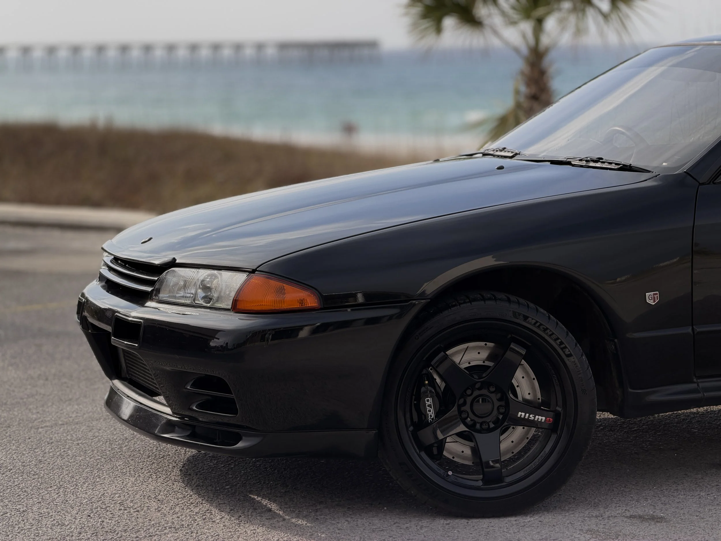 R32 NISSAN SKYLINE GTR FOR SALE
Close-up of the front part of a black sports car parked near a beach, with a palm tree and ocean in the background.