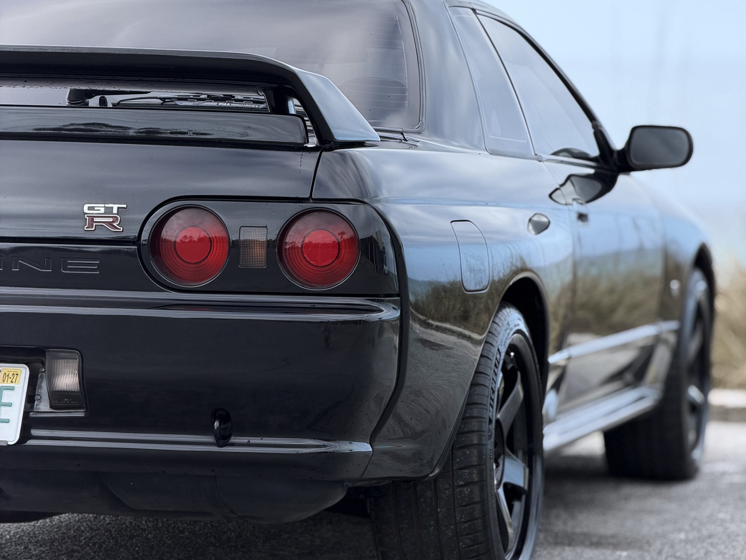 Close-up of the rear end of a black Nissan GT-R sports car, showing the taillights, logo, and rear wing, parked outdoors on a road or parking lot.